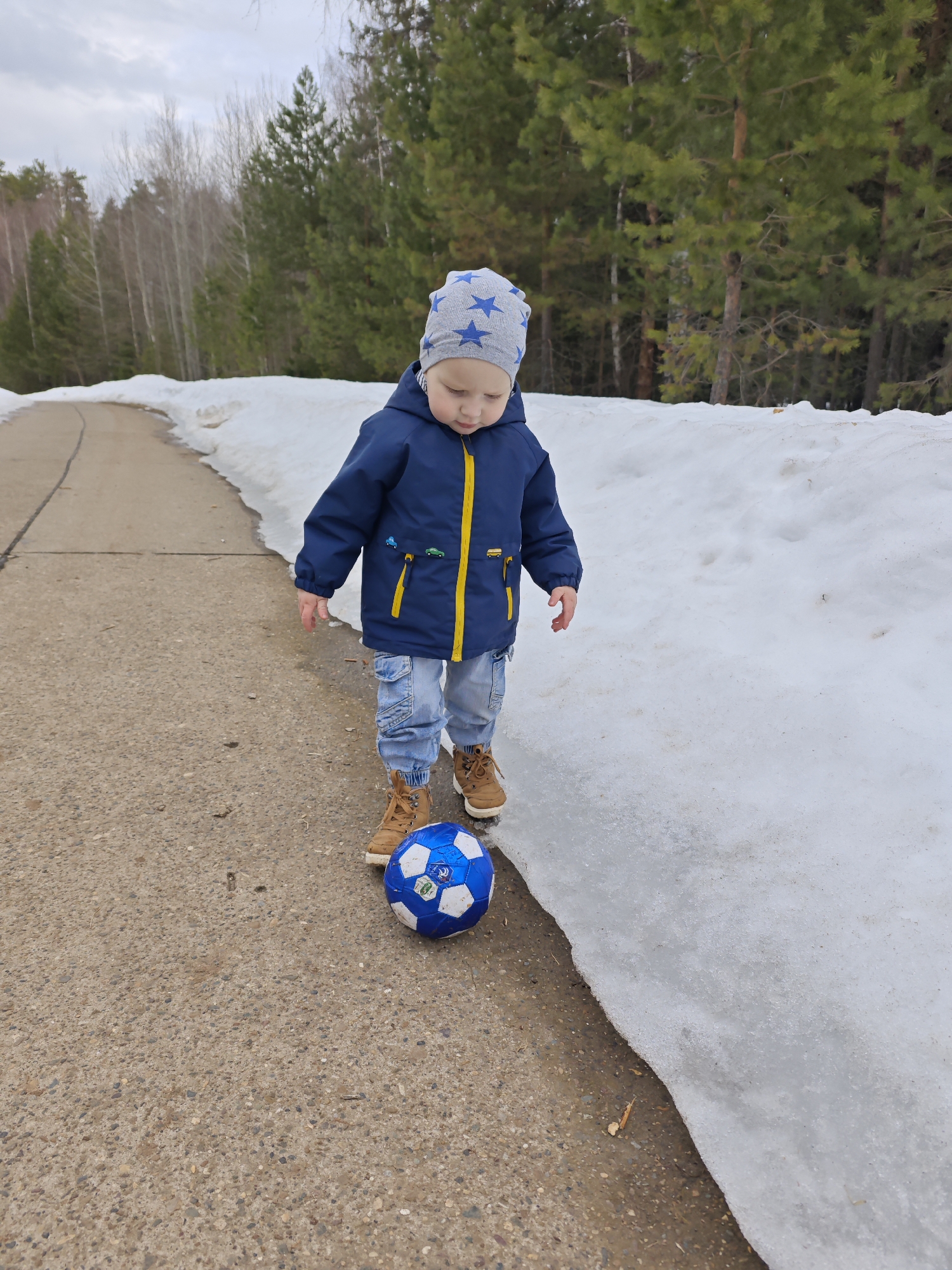 The image shows a child in Марковский, Russia, playing with a blue soccer ball on a paved path. The child is wearing a blue jacket with yellow trim, light-wash jeans, and brown boots. The child also wears a grey hat with blue stars. Patches of snow are visible along the side of the path, and trees line the background. The sky is overcast.