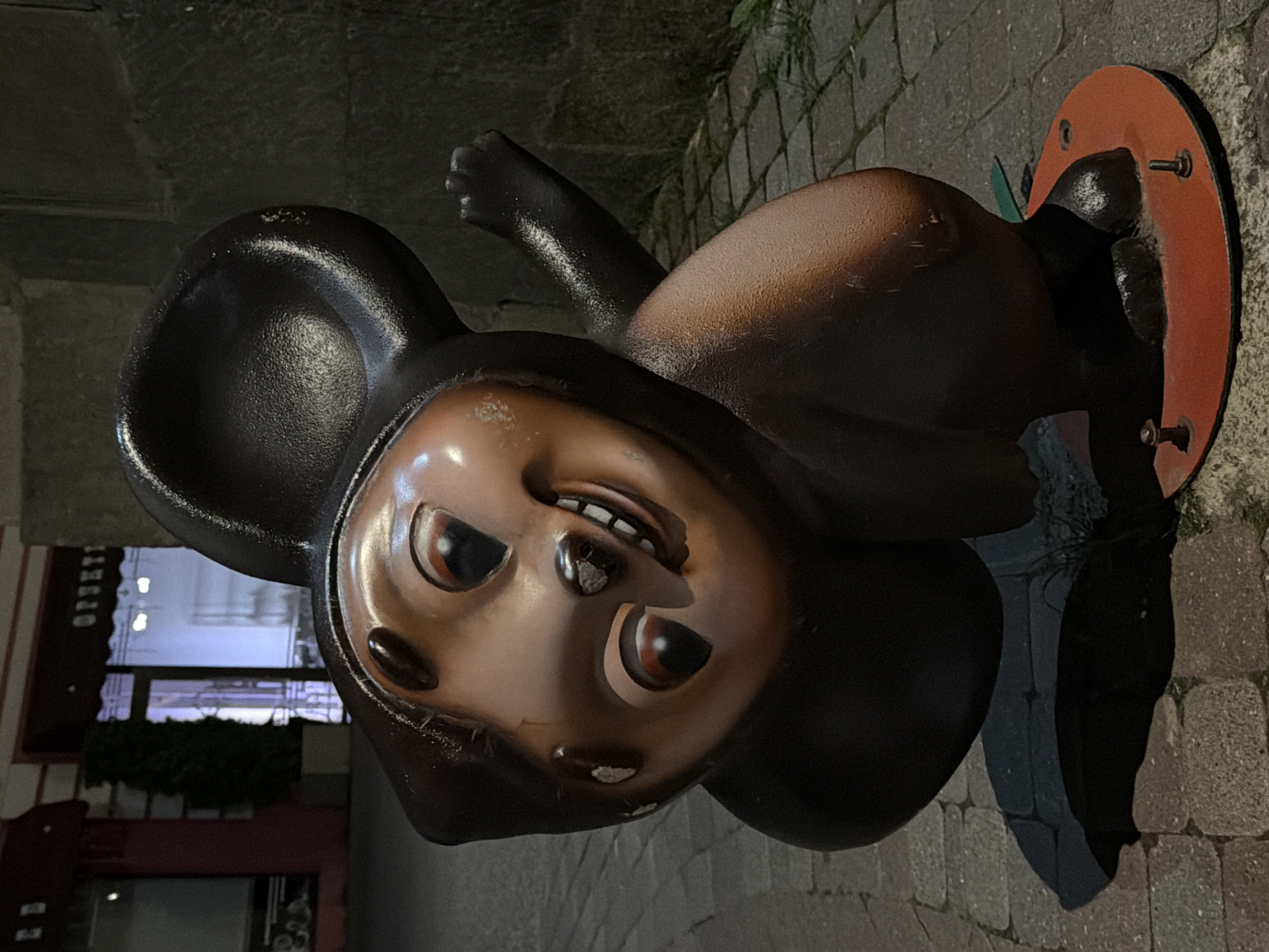 The image shows a statue of Cheburashka, a popular Soviet-era cartoon character, located in Kislovodsk, Russia. The statue is made of dark brown and bronze-colored material, depicting Cheburashka in a playful pose with a wide smile. It is situated on a cobblestone street, with buildings and shops visible in the background. The lighting suggests it is evening or night, with artificial lights illuminating the scene and casting shadows. The statue appears to be a popular landmark, as indicated by its prominent placement and the presence of a red circular base with bolts, suggesting it is a permanent fixture.