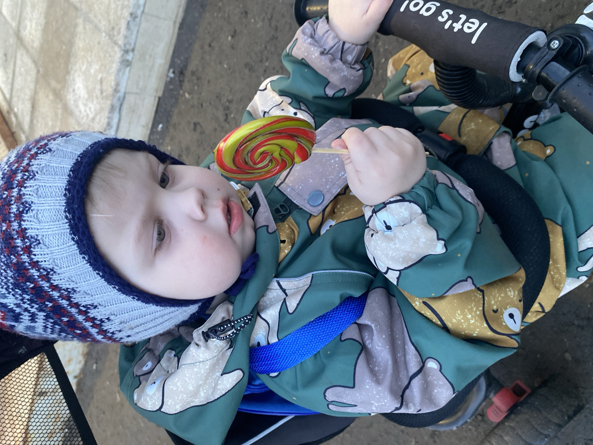 A young child wearing a blue, white, and red knit hat and a green and brown patterned jacket is sitting on a black stroller, holding a green and red lollipop. The handlebar of the stroller has "Let's go!" printed on it. The stroller is on a paved surface. The specific location in Chaikovsky, Russia is not discernable from the image.