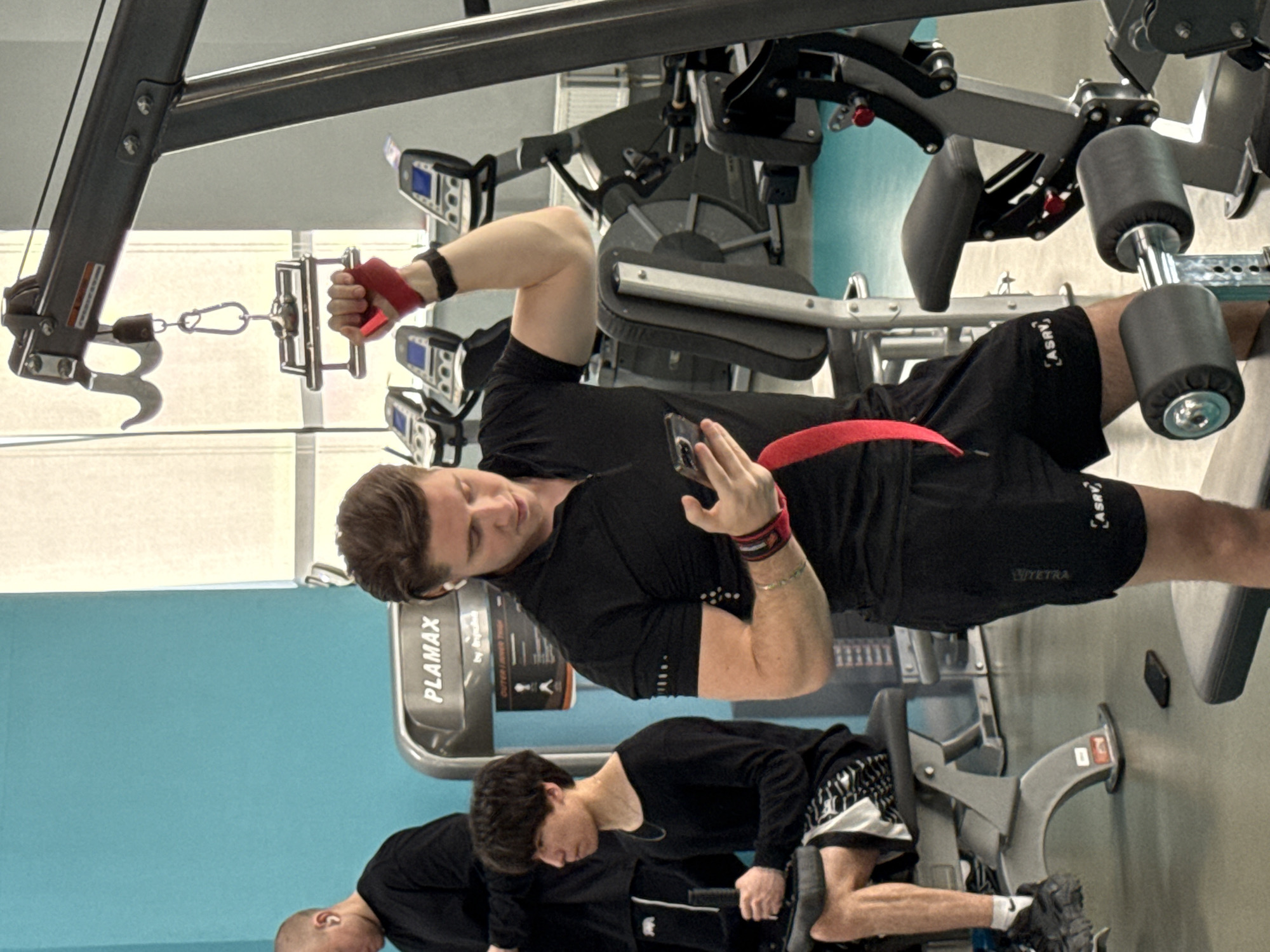 The image depicts an indoor gym or fitness center in Yekaterinburg, Russia, with several individuals engaged in exercise or rest. The primary subject is a man, appearing to be in his mid-twenties to early thirties, positioned centrally and looking down at a smartphone held in his left hand. He wears a black short-sleeved athletic top and black shorts. A red wrist wrap is visible on his right hand, which grasps a metal cable attachment from a multi-station gym machine. A red band or belt is also visible around his waist. His shorts feature the text "ASRV" on the left thigh and "VTYETRA" on the right.
The man is standing near a large, grey and black cable machine. To his right, partially visible, are the leg pads of another gym apparatus. In the background, two other men are present. One is seated on a gym machine, wearing a long-sleeved black top and patterned black and white shorts, with white socks bearing the "NIKE" logo. Another man's head is briefly visible on the far left. The machines feature small digital displays and red adjustment knobs. One machine in the background has "PLAMAX" written on its frame. The lighting suggests daytime. The overall atmosphere is that of a typical gym environment, with a light-colored floor and a light blue wall in the background providing a color contrast.