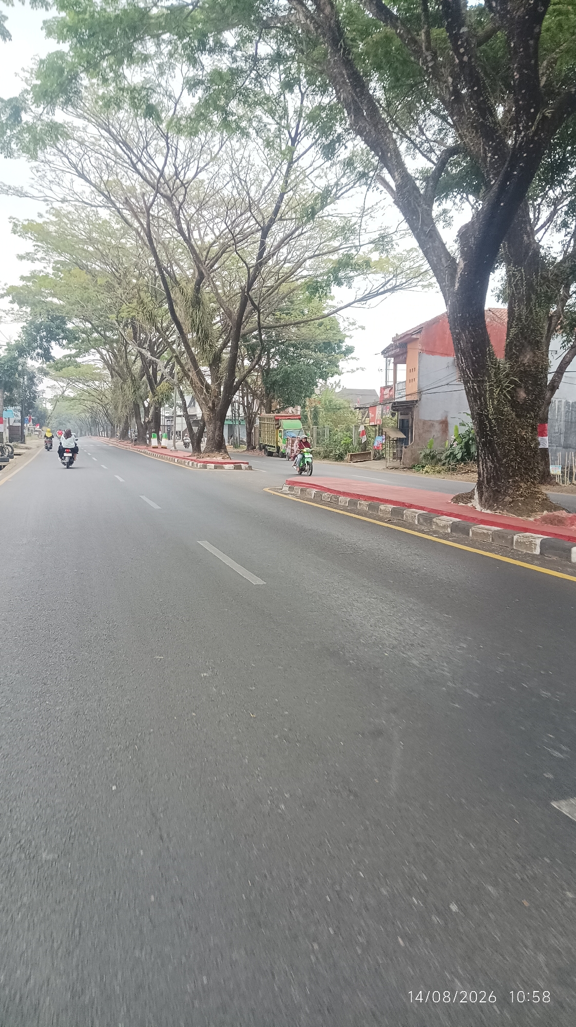 A long, gray asphalt road stretches into the distance, bordered by lush green trees on both sides. The road has white dashed lines marking the lanes. Motorcyclists are visible on the road, some riding and others stopped. Buildings line the right side of the road, with a red-tiled roof and orange-colored walls. A green truck is parked on the side of the road. Red and white concrete barriers separate the road from the sidewalk. The sky above is overcast. The timestamp "14/08/2026 10:58" is visible in the bottom right corner. This image appears to depict a scene in Tasikmalaya, Indonesia, likely a street within the city, given the presence of urban infrastructure, traffic, and residential/commercial buildings.
