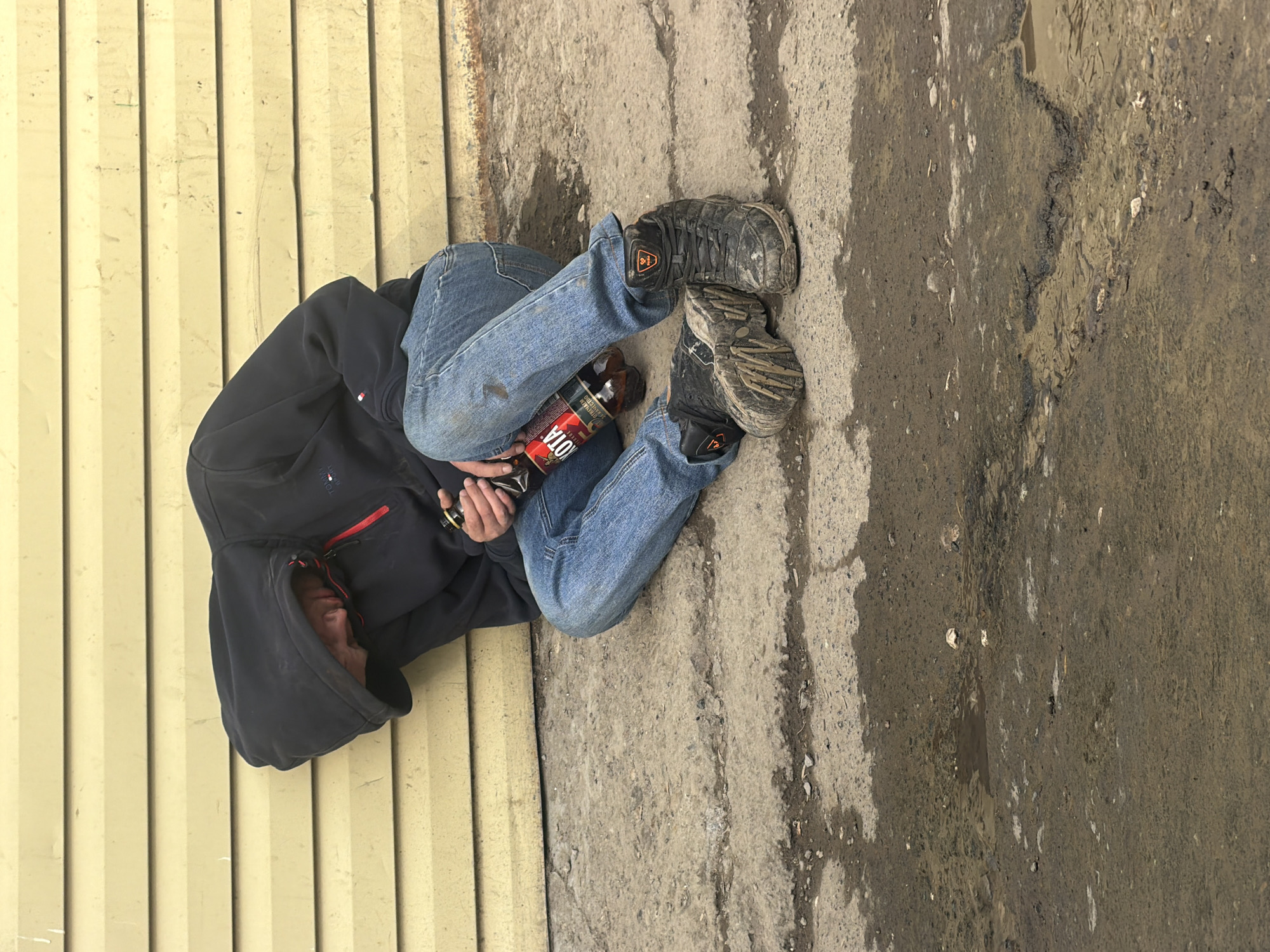 A person wearing blue jeans, a black hooded sweatshirt, and dirty work boots lies on the ground next to a yellow corrugated metal wall. The person is holding a bottle of "ОТА" brand alcohol. The ground consists of a mix of concrete and muddy patches. The photograph may have been taken in Perm, Russia, based on the bottle label.