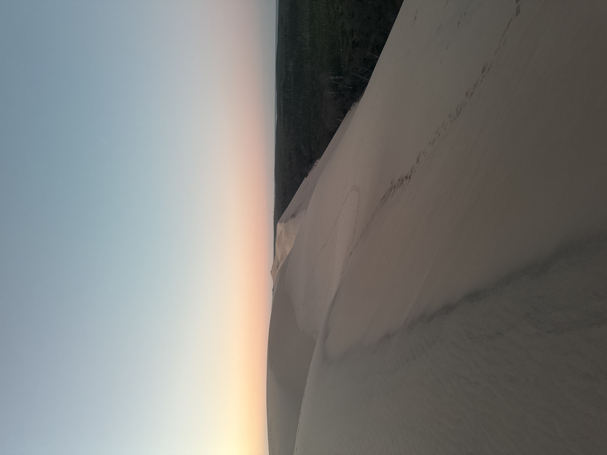 The image depicts the Dune of Pilat in La Teste-de-Buch, France, at sunset. The sky transitions from a pale blue at the top to soft hues of pink and orange near the horizon. A vast expanse of sand dunes dominates the foreground and midground, with gentle slopes and subtle shadows. Footprints are visible tracing paths across the sand. In the background, a dense, dark green forest stretches across the landscape, contrasting with the lighter tones of the dunes and sky. The overall scene is serene and natural, capturing the expansive beauty of the coastal dune environment.