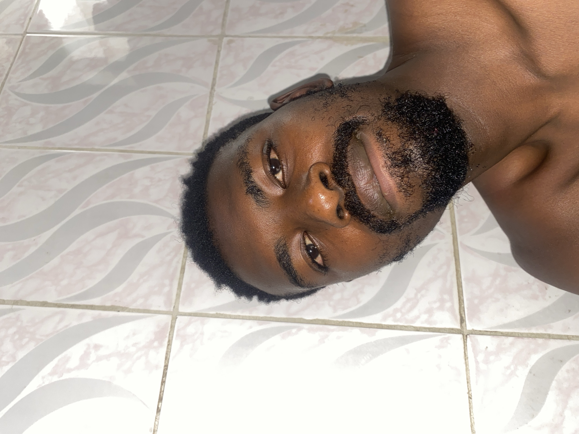 An adult Black man lies on his side on a tiled floor. The man has short, dark hair and a beard. The tiles are white with a faint grey wave pattern. No location in Barakin Akawo, Nigeria, is discernible from the image.