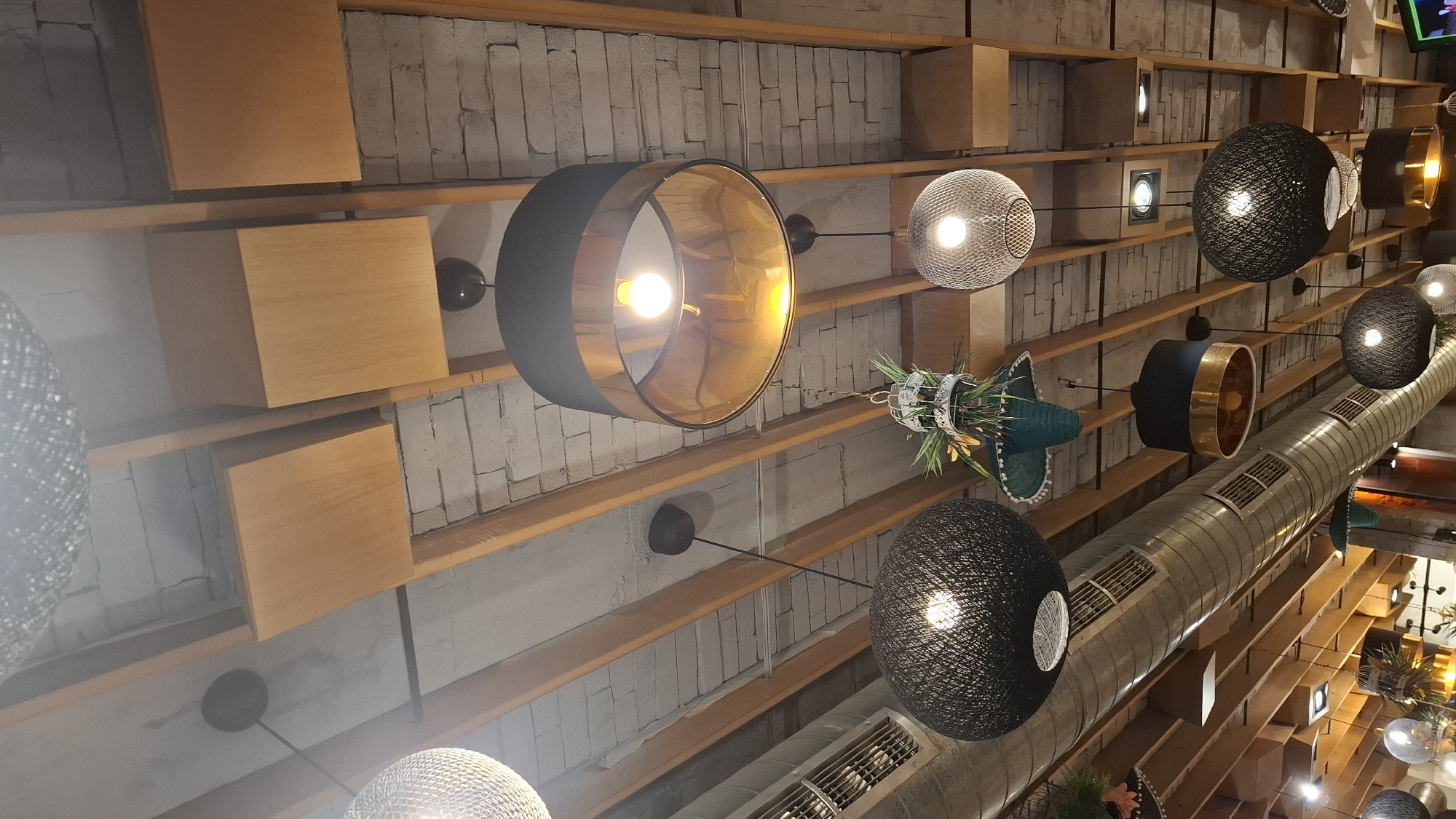 An interior view of a commercial establishment, likely a restaurant or cafe, located in Sofia, Bulgaria, is captured looking upwards towards the ceiling. The ceiling displays an architectural design featuring sections of exposed, light-colored brickwork interspersed with parallel wooden beams. Several flat, square wooden panels are integrated into the ceiling structure. A prominent, exposed cylindrical metallic ventilation duct equipped with multiple grilles extends horizontally across the upper-left quadrant of the image. A variety of pendant light fixtures hang from the ceiling. These include several large drum-shaped shades with black exteriors and reflective gold interiors, each housing an illuminated incandescent-style bulb. Multiple spherical pendant lights constructed from dark, woven string or rattan material, with visible glowing bulbs, are also present. One spherical pendant light made of a metallic mesh material is visible towards the right. Decorative elements include a white, birdcage-like hanging planter containing green foliage and artificial yellow-orange flowers, and a teal-colored, fringed hanging decoration also adorned with green foliage. Additional recessed spotlights are visible further within the ceiling framework. No individuals or specific actions are depicted within the frame.