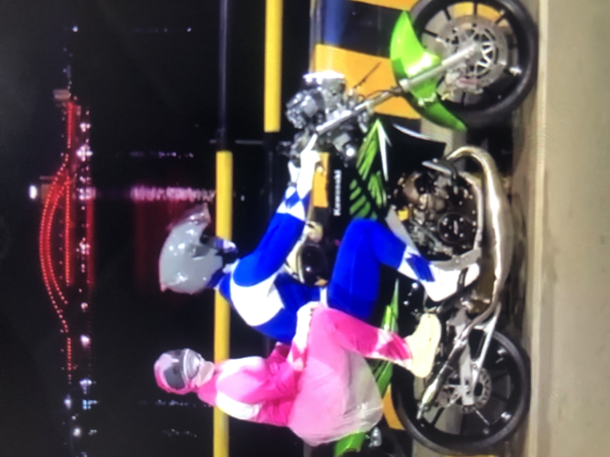 Two individuals dressed in Power Rangers costumes (one pink, one blue) are on a green and black motorcycle. The background shows a nighttime urban landscape with red lights, likely taken in Lhokseumawe, Indonesia. There are yellow and black striped barriers nearby. The Power Rangers are interacting with the motorcycle.