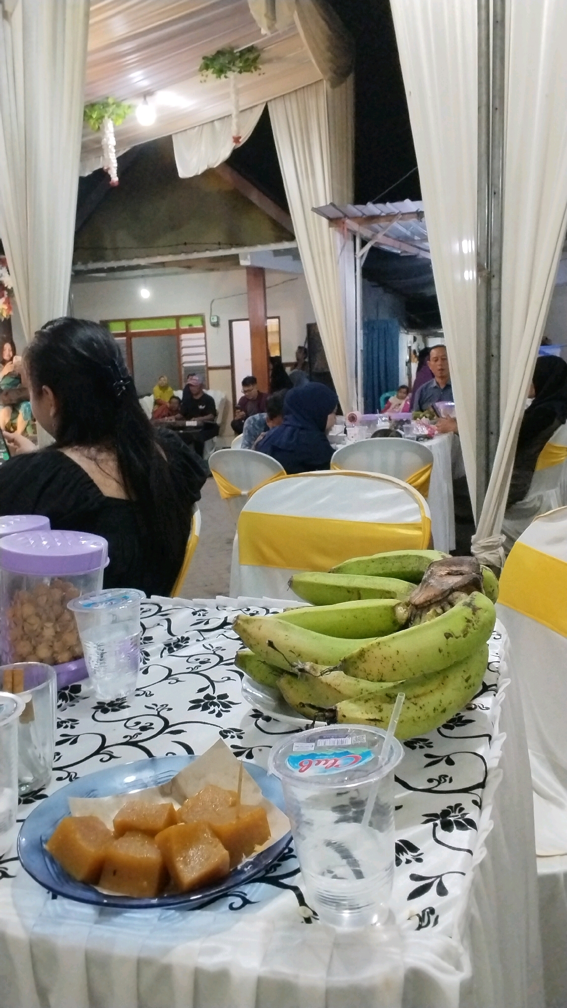 The image depicts an indoor gathering, likely a celebration or event, set in what appears to be a community hall or a decorated outdoor tent space in Penompo, Indonesia. The foreground features a table draped with a white tablecloth adorned with a black floral pattern. On the table are several items: a plate of cubed, golden-brown sweets, likely a traditional Indonesian dessert, accompanied by a toothpick; a clear plastic cup with a straw containing a clear liquid; and a bunch of green, unripe bananas. To the left, a clear plastic container filled with what seems to be snacks or nuts is visible, along with another clear cup.
In the mid-ground, more tables are set up, with chairs covered in white and yellow sashes. Several people are seated, some facing away from the camera, others in profile. One individual in the foreground, a woman with long black hair, is wearing a black top and is looking towards her left. Other attendees are visible in the background, some engaged in conversation or eating. The background suggests a ceiling with white draping, possibly a tent or a decorated indoor space, with lighting fixtures visible. The overall ambiance suggests a social event taking place in the evening.