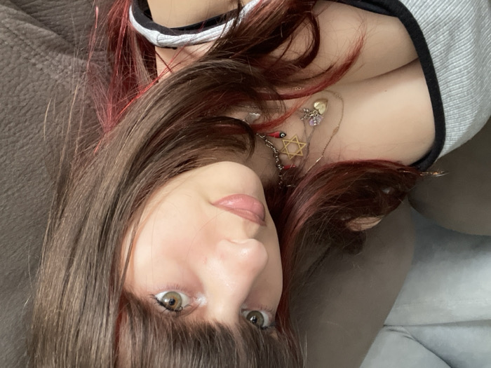 The image depicts a close-up, indoor portrait of a young female, observed from the chest up, within a private residence in Raanana, Israel. The subject has fair skin, long dark brown hair with red/pink highlights or tinsel, and bangs. Her eyes are light brown or hazel, and her lips have a glossy, light pink tint. She wears a light grey ribbed tank top with black trim along the neckline and straps. Visible on her neck and chest are multiple necklaces, including a silver chain with a small golden heart pendant and a purple charm, and a red choker-style necklace featuring a golden Star of David charm and a small red evil eye bead. The background shows a portion of a brown, upholstered surface, consistent with a sofa or armchair. The subject is looking directly towards the camera with a neutral expression.