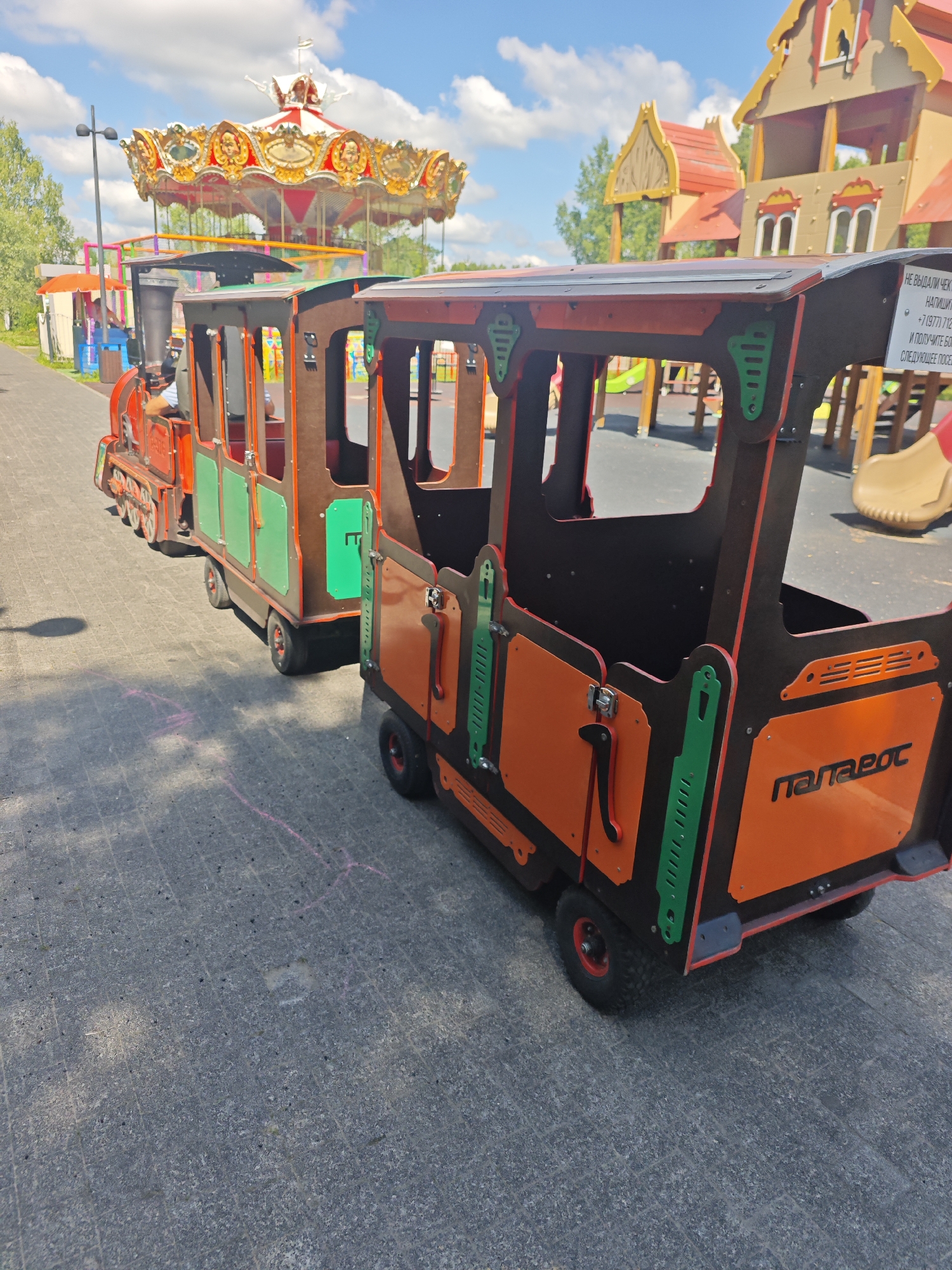 The image shows an amusement park with a carousel and a toy train. The toy train is made of wood and metal, with orange, green, and brown colors. It has two carriages and appears to be on display or for rent. A sign on the train reads "Папаеот" (Papaet) in Russian. In the background, a carousel with a red and gold canopy is visible, along with a children's playground with wooden structures. The ground is paved with grey tiles. The sky is blue with scattered clouds. The location is identified as an amusement park in the city of Chekhov, Russia.