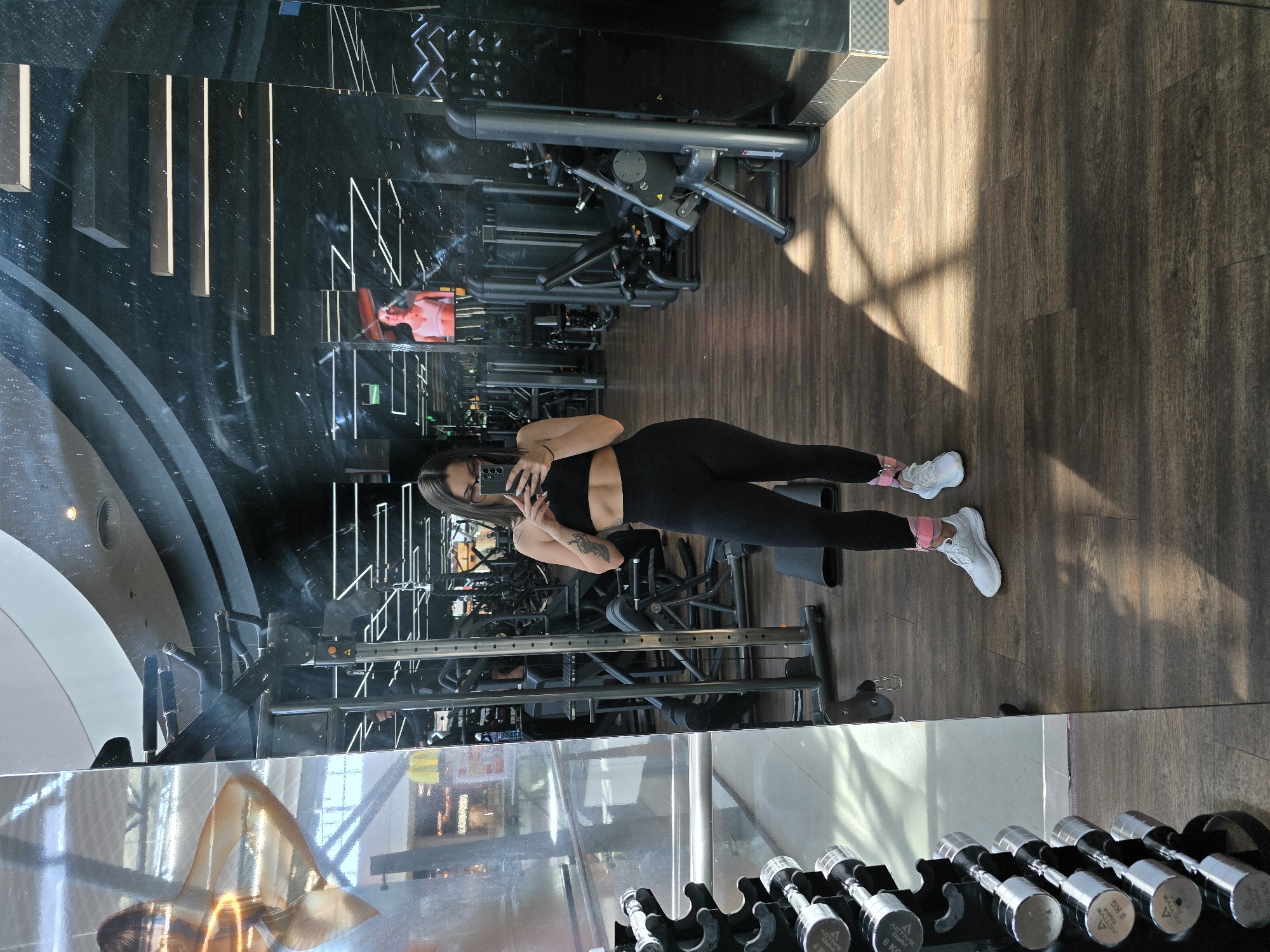 A young woman is captured in a mirror selfie taken within a modern gym setting in Burgas, Bulgaria. She stands centrally in the frame, holding a smartphone to photograph her reflection. She is dressed in black athletic wear, comprising a sleeveless crop top that reveals her midriff and form-fitting leggings. Her footwear consists of white sneakers accented with bright pink ankle wraps or socks. A tattoo is visible on her left arm.
The gym environment is reflected extensively in the large mirrors that dominate the scene. Various gym equipment, including weight machines and benches, are visible in the background reflections. The floor is made of dark wooden parquet. In the lower foreground, a rack of chrome dumbbells is clearly visible, with some bearing the brand name "ACTIVE" and weight markings like "4 KG," "6 KG," and "8 KG."
Natural light streams into the gym, creating distinct shadows on the wooden floor, suggesting the picture was taken during daytime. Reflections in different sections of the mirror also show abstract architectural elements, possibly lighting fixtures, and glimpses of other gym patrons, including one person wearing a red top. The lower left portion of the mirror reflects an artistic image of a woman, possibly a decorative advertisement. The overall scene portrays a typical, well-equipped fitness facility.