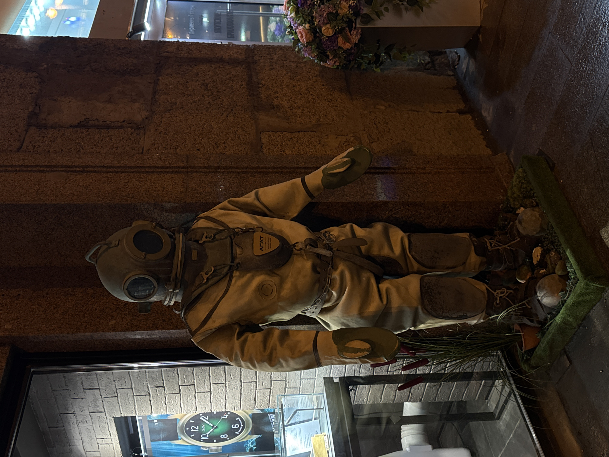 A full-bodied, old-fashioned diving suit, likely a mannequin, lies horizontally in a display. The suit is beige with reinforced knee and elbow areas. The helmet is a spherical shape with round glass windows. It has a tag that reads "ATAT". The suit rests on a bed of rocks and artificial grass, with some planted decorative grass. The display is next to a brick wall, and above it is a flower arrangement. The bottom left corner of the image shows a glass case with a clock display. The location is Moscow, Russia, based on an inscription in Cyrillic lettering above the diving suit.