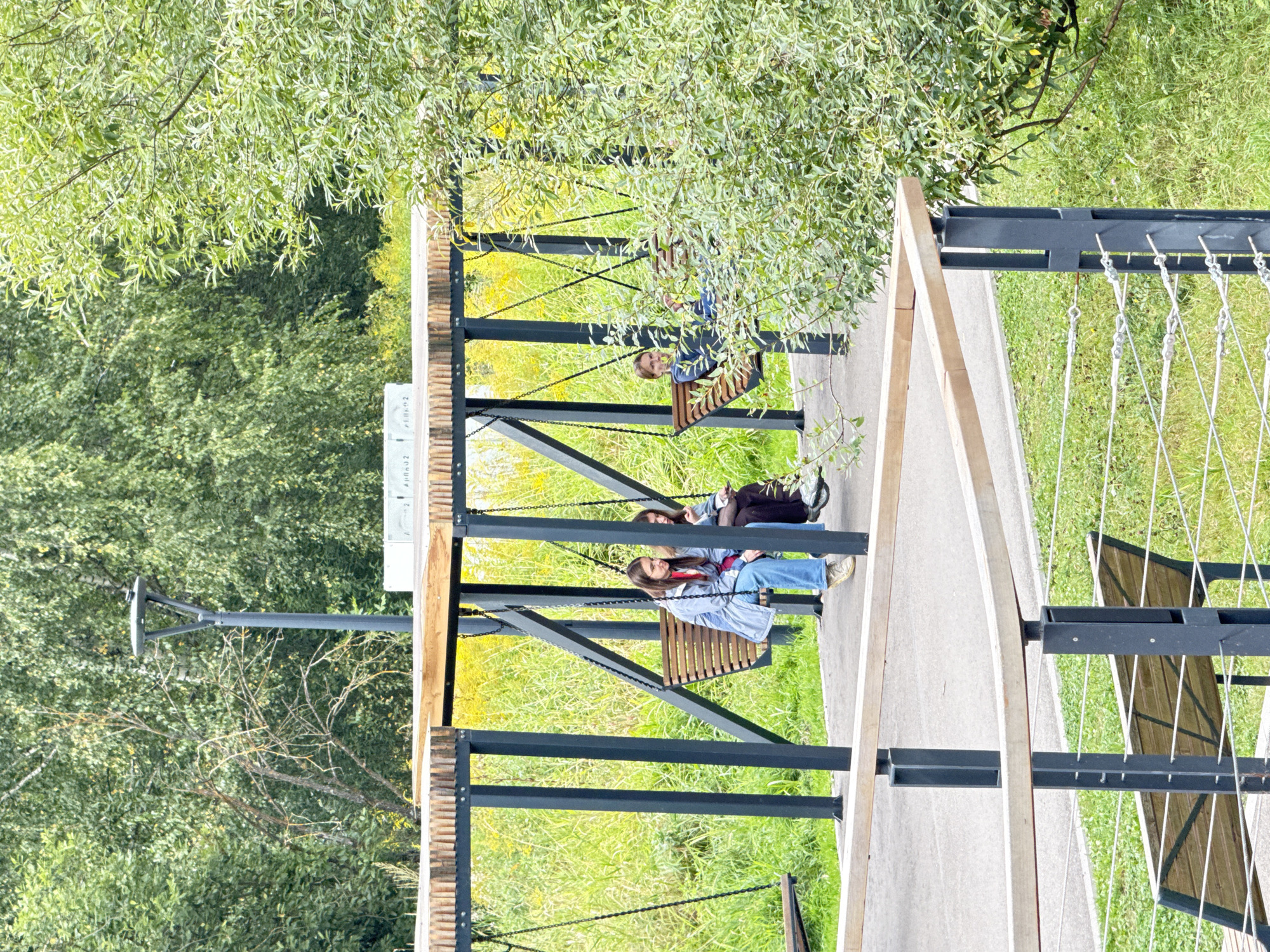 The image depicts an outdoor public park area, likely in Moscow, Russia, featuring a modern swing set structure with a wooden roof supported by dark metal beams. Three individuals are visible on two of the swings. On the left swing, two young women are seated; one in a light blue jacket and jeans, the other in darker clothing. On the right swing, a young person is seated, facing the camera. They are surrounded by lush green grass, tall reeds, and trees, suggesting a natural, undeveloped landscape within the city. The lighting indicates it is daytime, possibly overcast given the diffused light.