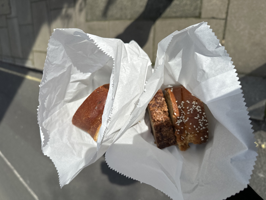 Two open white paper bags containing baked goods are presented in an outdoor setting, likely a street or pavement in the city of London, United Kingdom. The upper bag contains a single, reddish-brown, somewhat glossy baked item, possibly a bun or bread piece. The lower bag holds two distinct baked items: an upper, darker brown slice with a coarse, crumbly top, and a lower, lighter brown slice topped with white sesame seeds. The items are viewed against a backdrop of dark grey asphalt or tarmac with a subtle yellow line, bordering lighter grey paving stones. Strong, directional sunlight from the left casts defined, dark shadows of the bags and an unseen object onto the ground, indicating a bright, clear day. No individuals are visible.
