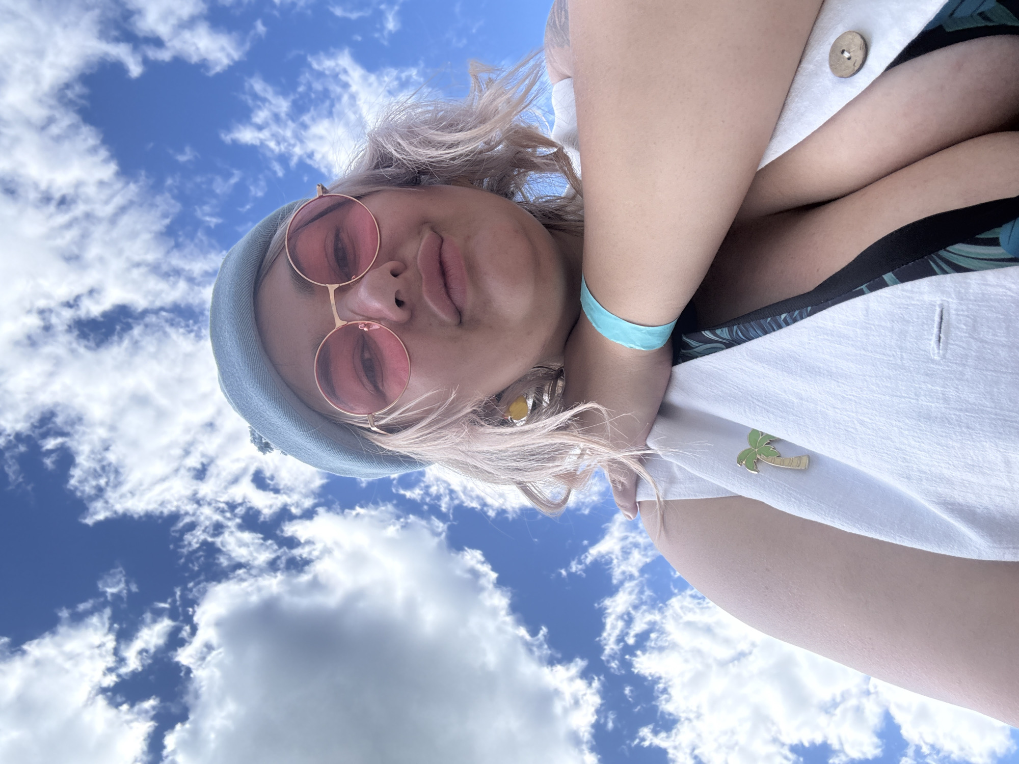 A close-up, low-angle shot shows a person outdoors against a bright blue sky with scattered white clouds. The person is wearing a light blue beret, pink-rimmed sunglasses, and a white vest over a patterned top. A turquoise wristband is visible on their left arm. A small, gold palm tree pin is attached to the vest. The person's head is tilted back, and they are looking towards the camera. The background is entirely sky.