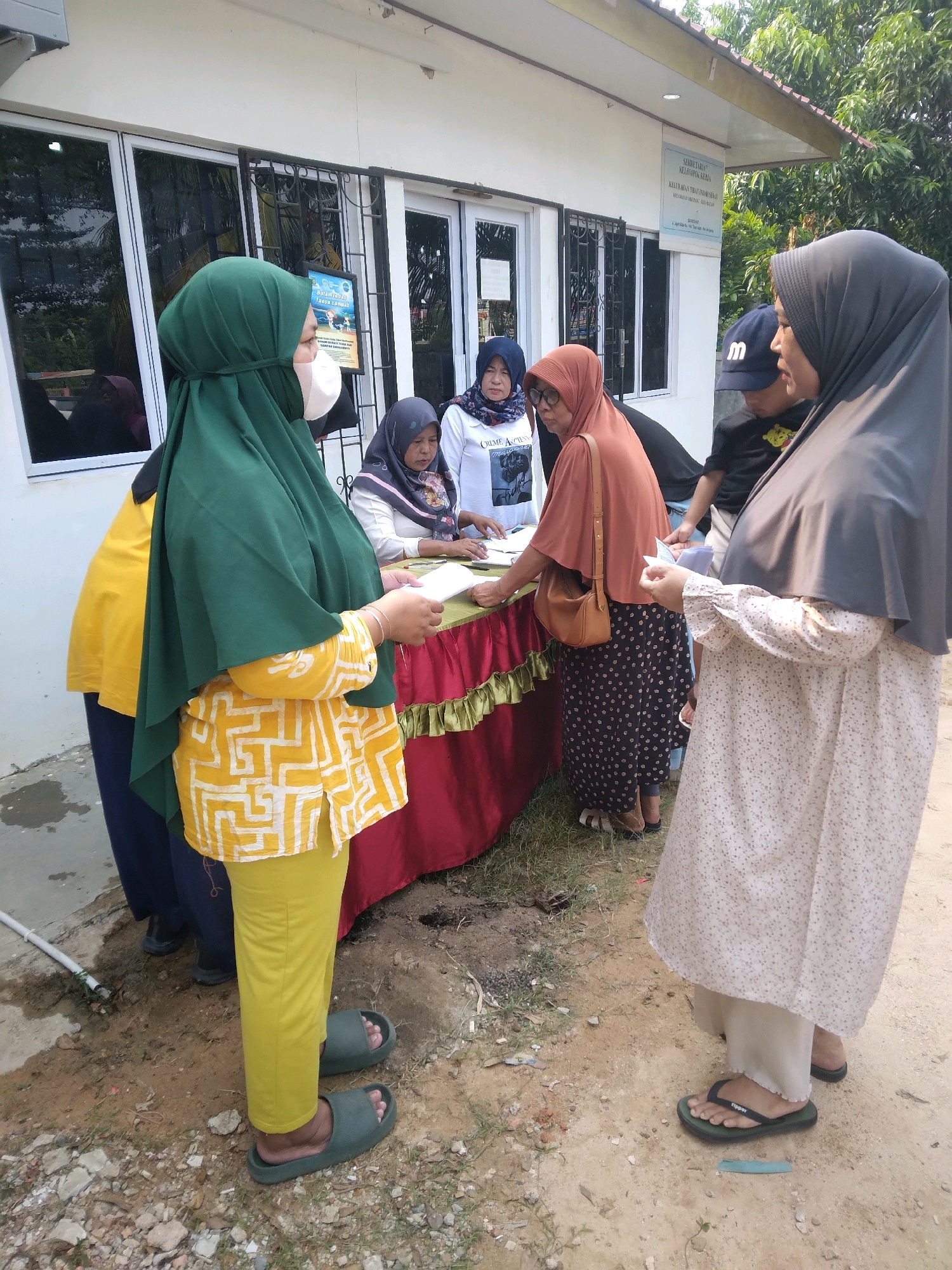 In Batam, Indonesia, a group of people, primarily women wearing hijabs, are gathered outside a building. The building appears to be a government or community office, indicated by the signage on the wall that reads "SEKRETARIAT KELURAHAN LABUH RAH". A red and green draped table is set up, with papers and brochures on it. Several individuals are interacting around the table, with some appearing to look at or handle documents. The environment is outdoors, with a sandy and gravelly ground surface, and vegetation visible in the background. One woman in the foreground wears a green hijab and a yellow patterned top, while another to her right is dressed in a light-colored floral dress and a dark hijab. A person in the background wears a navy blue baseball cap with an "M" logo. The scene suggests a community event or an administrative process taking place in an open area adjacent to the building.