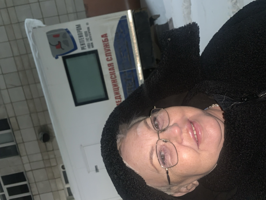A middle-aged to elderly woman with light hair and gold-framed eyeglasses is prominently featured in the foreground, looking directly at the camera with a slight smile. She is wearing a black, textured hooded jacket. Behind her, a white mobile medical unit, likely a truck or trailer, is parked on snow-covered ground. The vehicle's side displays Cyrillic text in red, including "РЕНТГЕНОРOM" (RENTGENOROM) and "МЕДИЦИНСКАЯ СЛУЖБА" (Medical Service), along with a logo depicting a stylized medical imaging device. A dark, rectangular window or screen is visible on the medical unit's side. In the upper background, a multi-story building with a light-colored, possibly tiled or bricked, facade and multiple windows is present. The ground is covered in snow. This scene is situated in Chaikovsky, Russia.