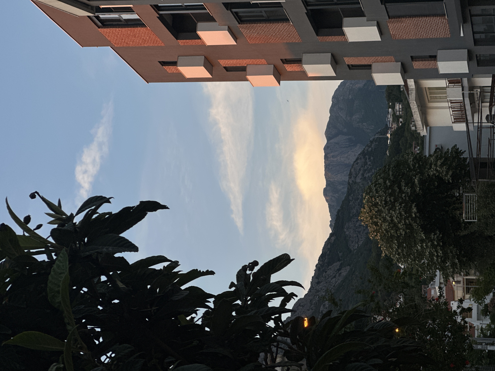 The image depicts a view from Šušanj, Montenegro, framed by two buildings at the top. The building on the left has a reddish-brown brick facade and projecting square balconies, while the building on the right is gray with white balconies. The sky is visible between the buildings, with scattered clouds and a faint yellow hue near the horizon, suggesting either sunrise or sunset. A mountain range forms the background. Dark, leafy trees occupy the lower portion of the frame, obscuring parts of a town below.