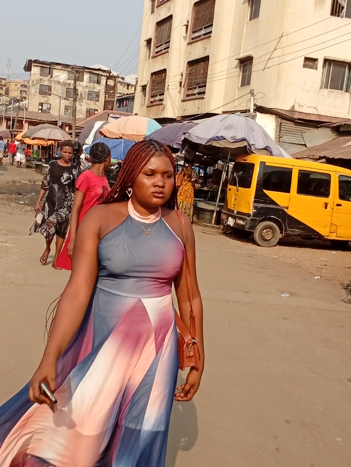 A woman with brown braided hair, wearing a flowing halter-neck dress, walks down a crowded city street. The dress is a blend of gray, pink, and blue. She carries a small brown purse on her shoulder and holds a phone in her hand. Behind her, people walk amongst market stalls with colorful umbrellas and a yellow taxi van. Buildings are visible in the background, adding to the urban scene.
