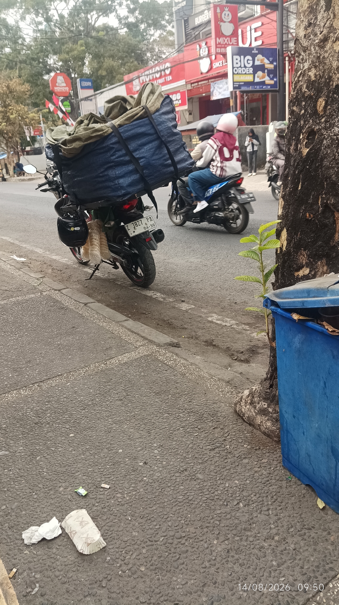 In Tasikmalaya, Indonesia, an outdoor scene captured on August 14, 2026, at 09:50, depicts a street with several motorcycles and commercial buildings. A motorcycle with a large, blue tarp-covered load is parked on the right side of the road. Another motorcycle is being ridden by two individuals, a driver and a passenger wearing helmets, one with a pink head covering. In the background, commercial establishments with red signage are visible, including one with the name "MIXUE" and another advertising "BIG ORDER." A blue trash bin stands near a tree on the right side of the image. The street is paved with asphalt and cobblestones, and there is some litter on the ground.