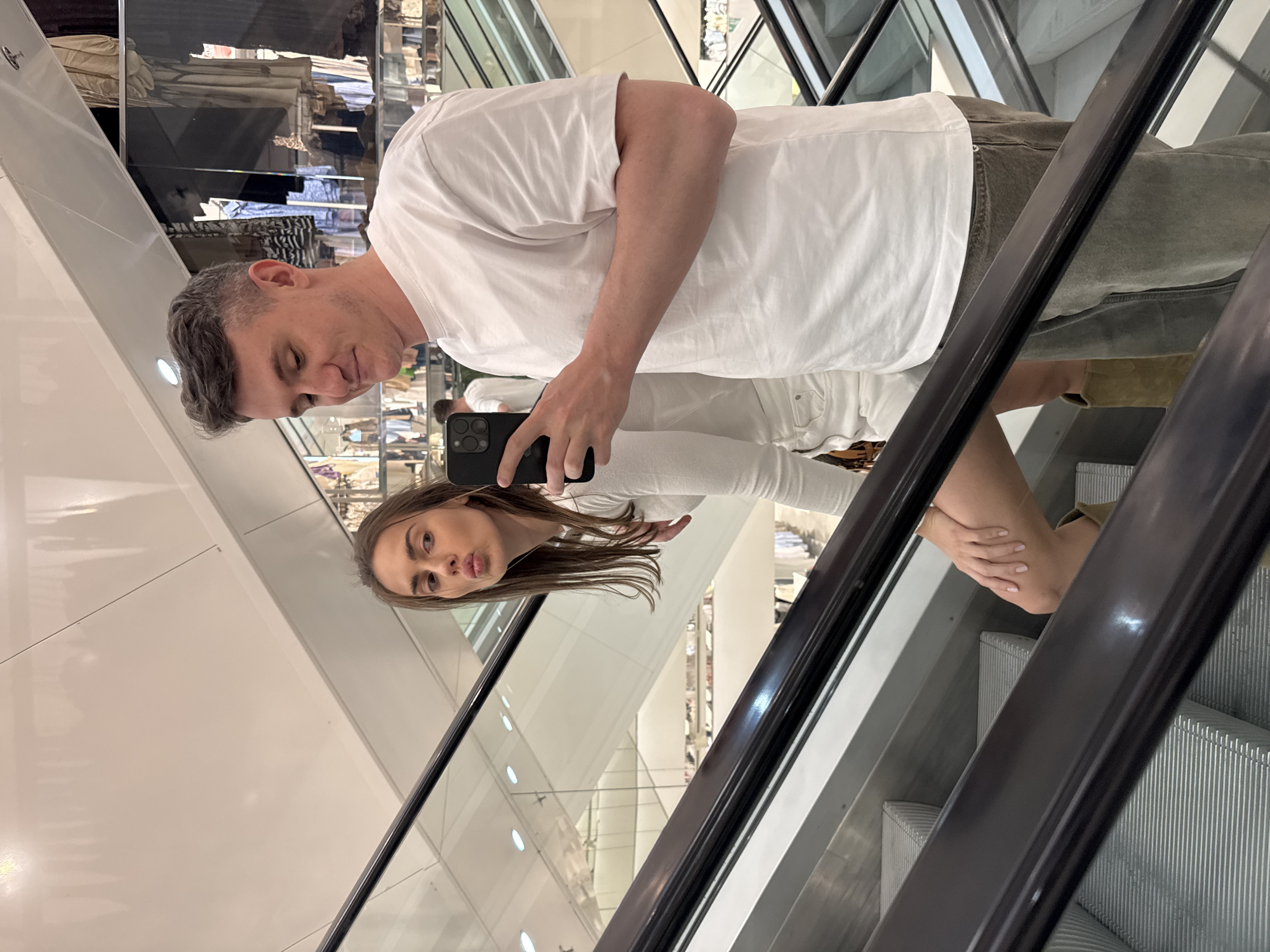 A man and a woman take a selfie on an escalator. The man has short, gray hair, a white t-shirt, and olive green pants. The woman is wearing a long-sleeved white shirt. They are in a clothing store, as clothing racks are visible in the background.