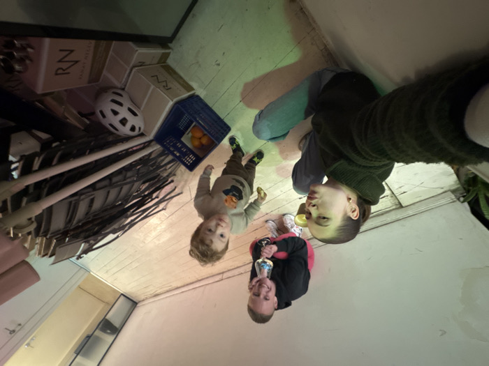 The image depicts an indoor scene within Tel-Aviv, Israel, featuring three individuals on a light-colored wooden floor. A female subject in the foreground, wearing a dark green sweater and light blue jeans, is positioned to take a selfie, puckering her lips towards the camera. To her right and slightly behind, a second adult female crouches, holding a stemmed glass containing a light-colored liquid. She wears a black hoodie, pink pants, and white athletic shoes, displaying a smile. A young male child stands between the two women, looking towards the camera with a slightly open mouth, holding a small yellow object. He is dressed in a light-colored long-sleeve shirt with a graphic and brown pants. The background consists of plain light-colored walls, with a stack of dark-colored folding chairs leaning against the right wall. On the floor, near the child, are a blue plastic crate holding three oranges, several beige cardboard boxes marked 'RN', and a white bicycle helmet. A yellow cup is visible on the floor near the crouching woman. The room's appearance suggests a casual, possibly utilitarian or multi-purpose space.