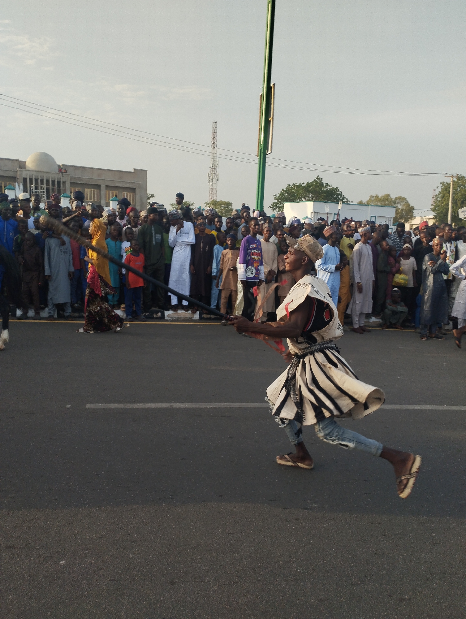 The image depicts a man running on a street in Gombe, Nigeria, holding a long pole. He is wearing a traditional costume with a striped skirt, a hat, and sandals. A large crowd of people watches from behind a barrier along the roadside. A building with a dome is visible in the background, along with a telecommunications tower. A street light pole also stands near the crowd. The sky is overcast.