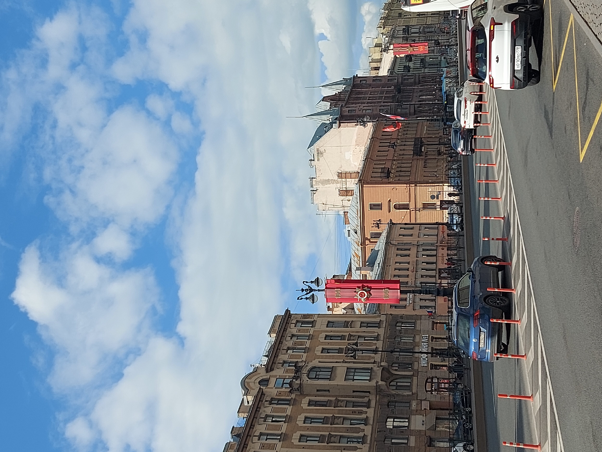 The image depicts a street scene in Saint Petersburg, Russia. Several buildings line the street. Red banners are seen along the street. Vehicles are parked along the side of the road, separated from the traffic lane by rows of orange bollards. A partly cloudy blue sky is visible above the buildings.