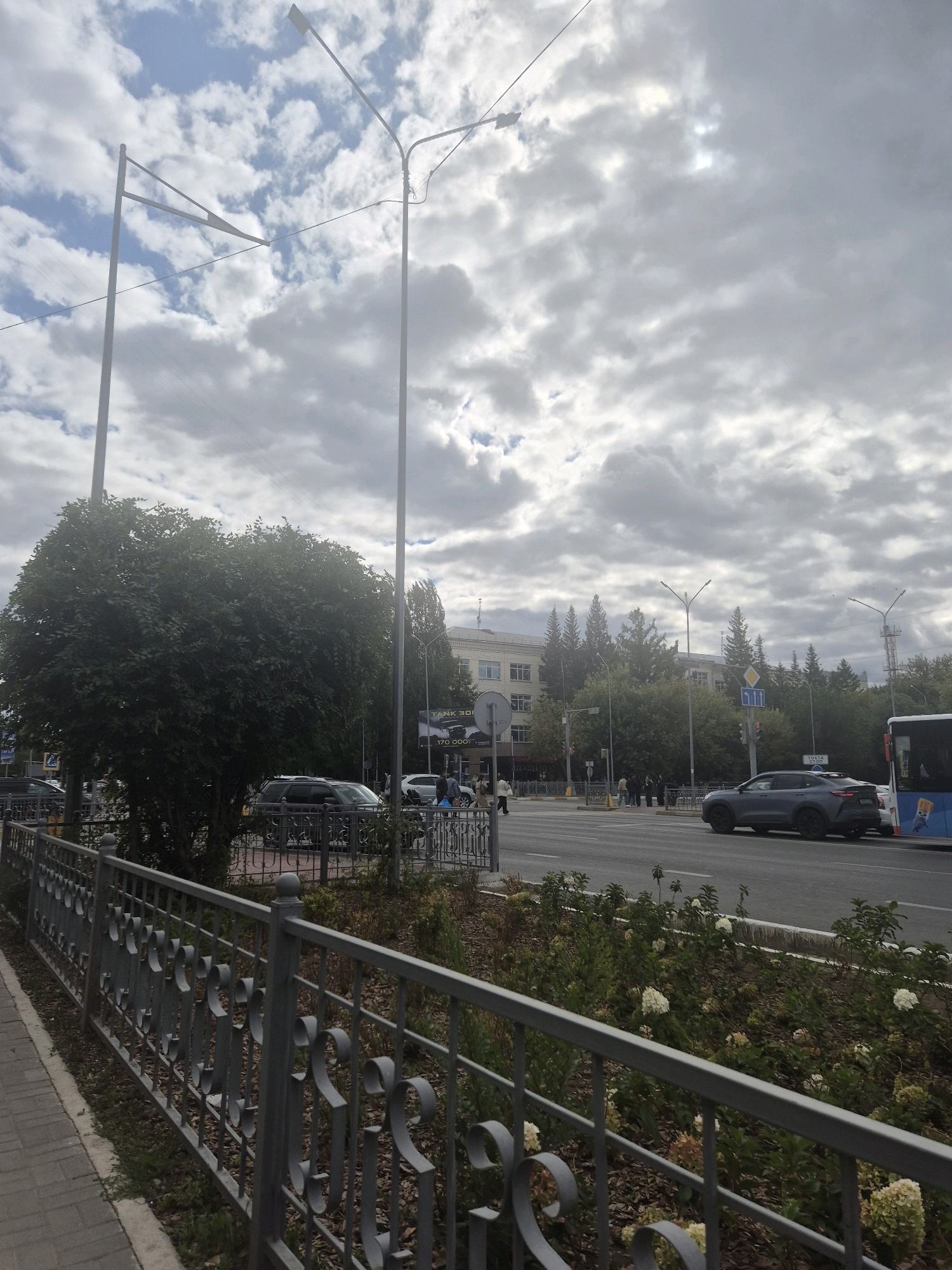 The image captures a view of a city street in Kokshetau, Kazakhstan. The foreground features a gray decorative metal fence and a paved sidewalk. Beyond the fence, a road is visible with several cars, including a dark gray sedan and a white car. A large, dark green tree stands to the left of the road. In the background, a multi-story building with a light-colored facade and numerous windows is present, surrounded by trees. Two streetlights are prominent in the sky, which is filled with clouds. A bus is seen on the right side of the road. Several pedestrians are walking on the sidewalk in the distance. The overall impression is an urban street scene under a cloudy sky.