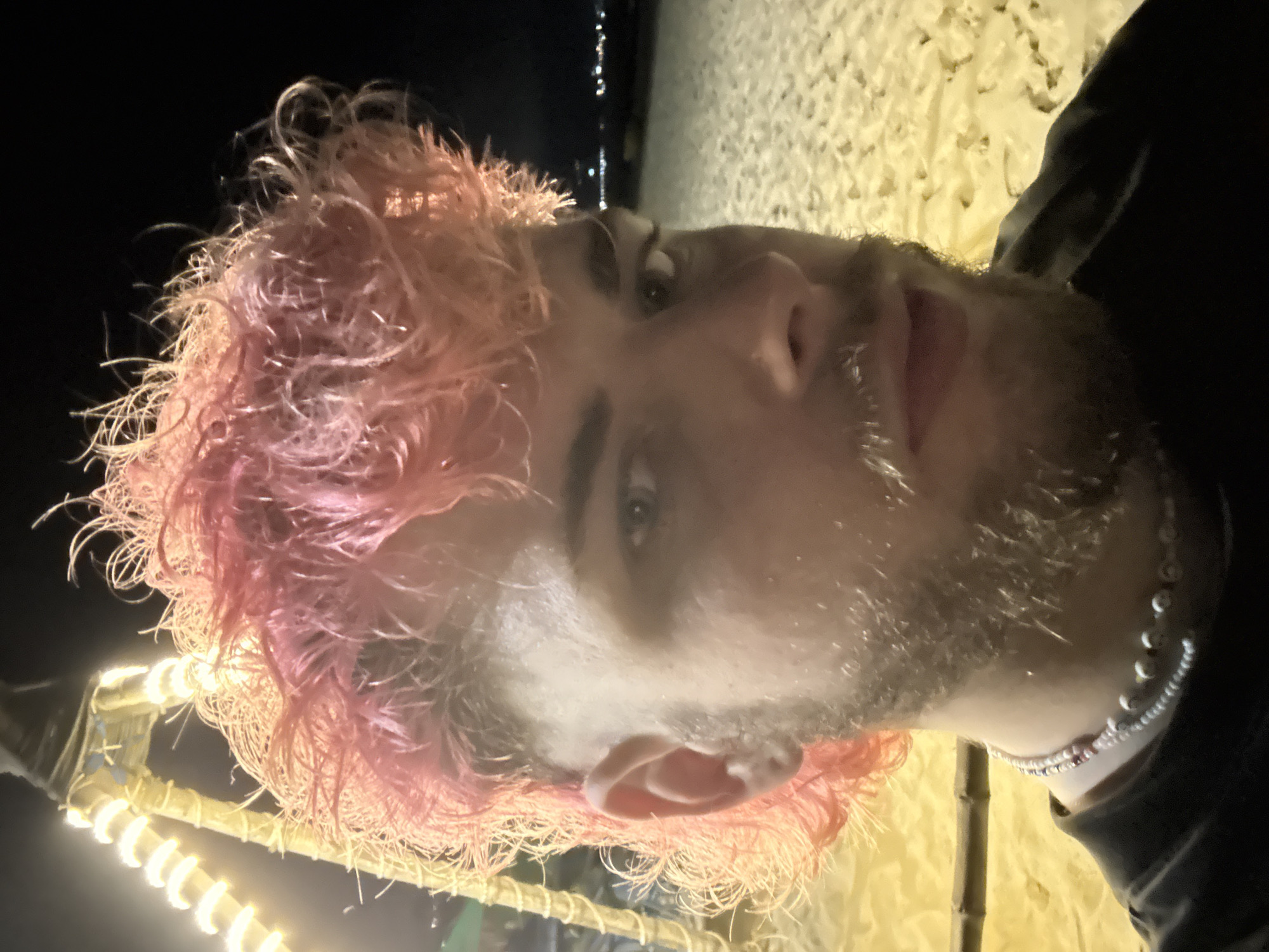 The image is a close-up, low-angle portrait of a man with pink hair and a short, light-colored beard. He is wearing a beaded necklace. The background shows a dimly lit beach with lights in the distance, potentially indicating a city skyline. This scene is likely in Đà Nẵng, Vietnam. A string of yellow lights is visible in the lower left corner of the image.