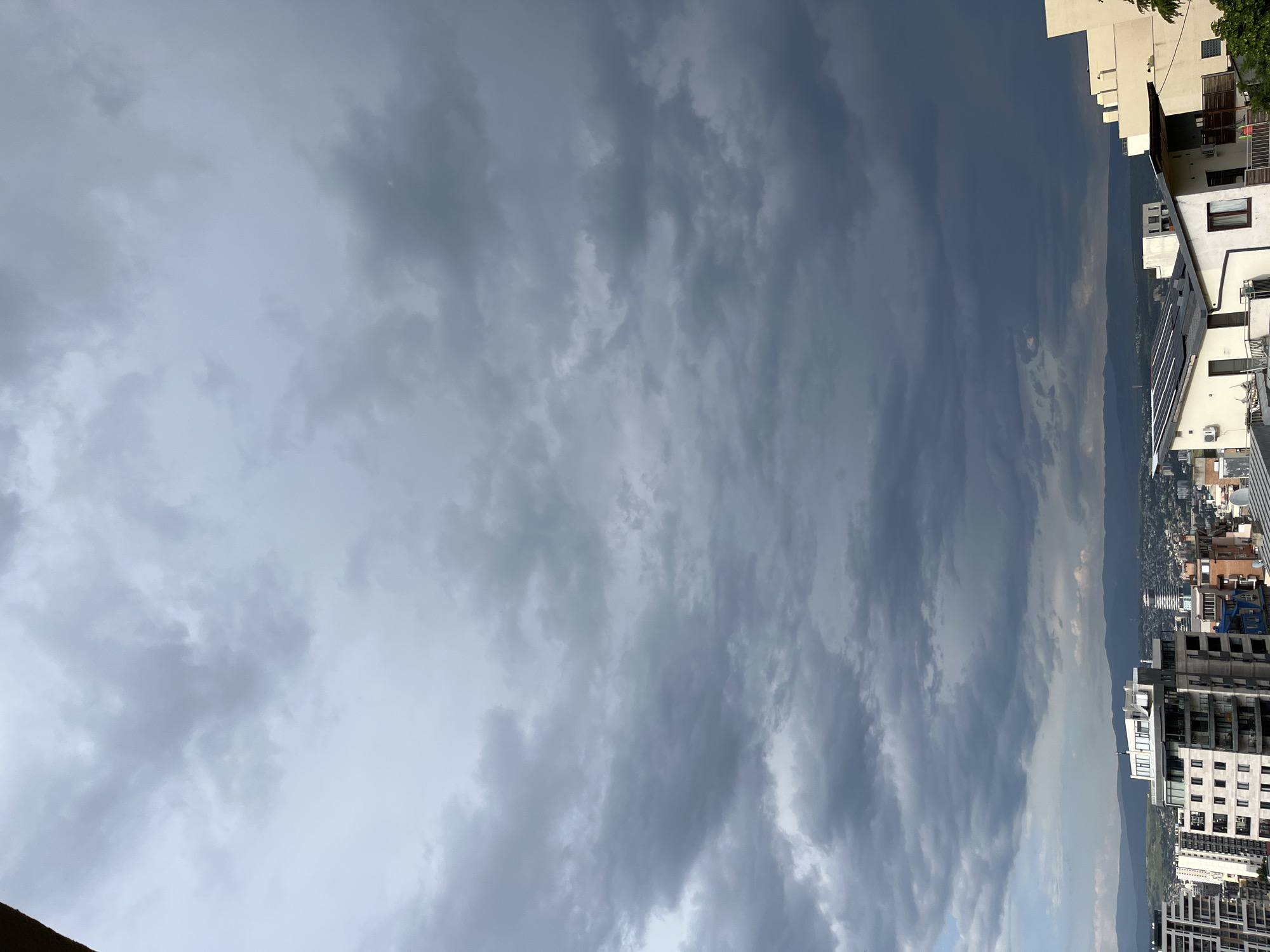 The image shows a cityscape under a cloudy sky. The sky is filled with gray clouds, indicating potential rain or stormy weather. Below the sky, there is a view of a city with buildings of various sizes and designs. Some of the buildings appear to be residential, while others may be commercial or office structures. In the distance, there is a suggestion of mountains, adding depth to the scene. The angle of the photograph is rotated. The buildings appear on the right side and the clouds are on the left.