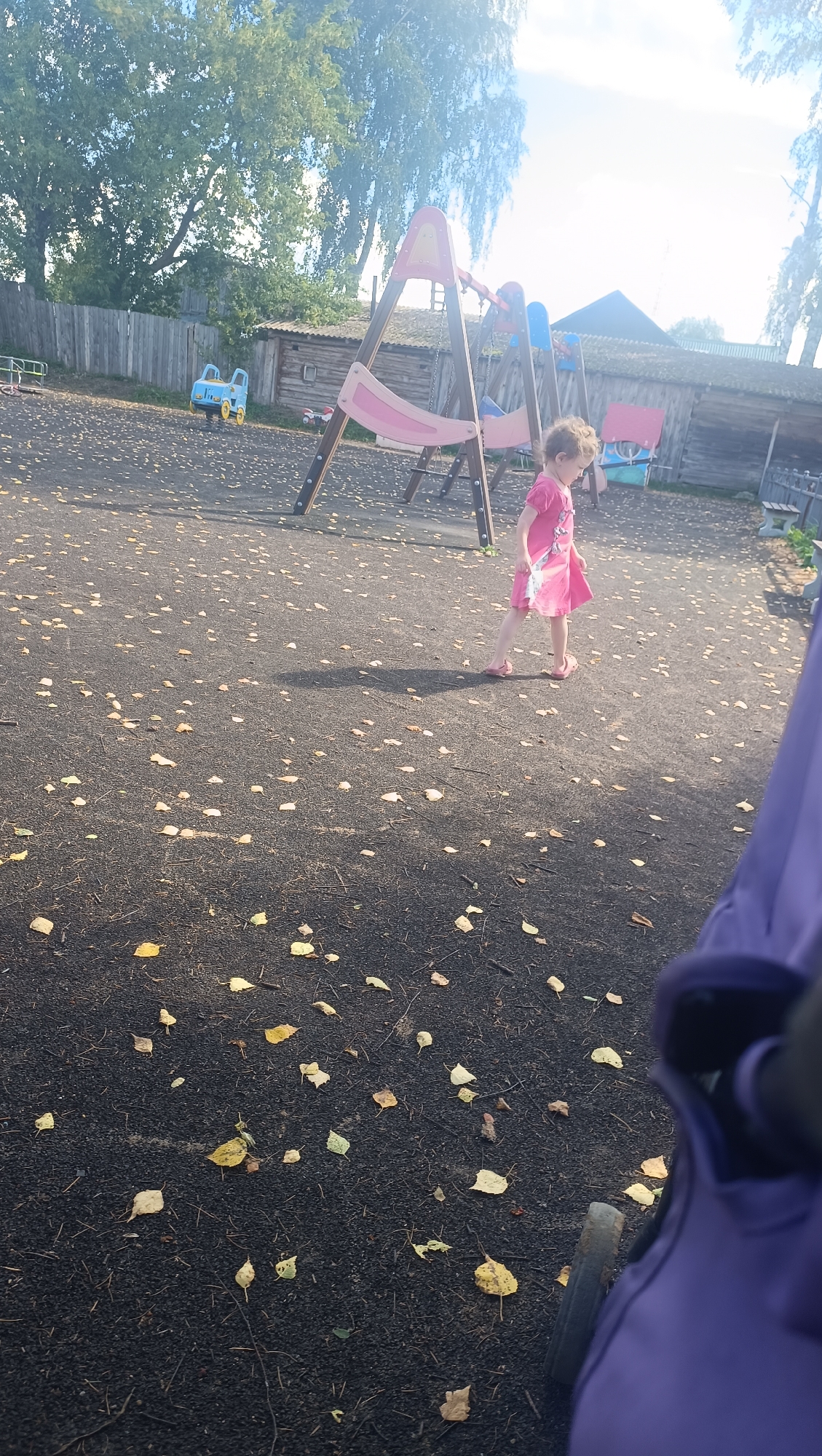 In Karsha, Russia, a young child, a girl wearing a pink dress, walks across a dark, leaf-strewn ground. The child is near a playground with a swing set that features pink and blue elements. To the left of the swing set, a small blue toy vehicle is visible. In the background, wooden structures and a fence are present, with trees and a bright sky overhead. The perspective suggests the image was taken from a stroller.