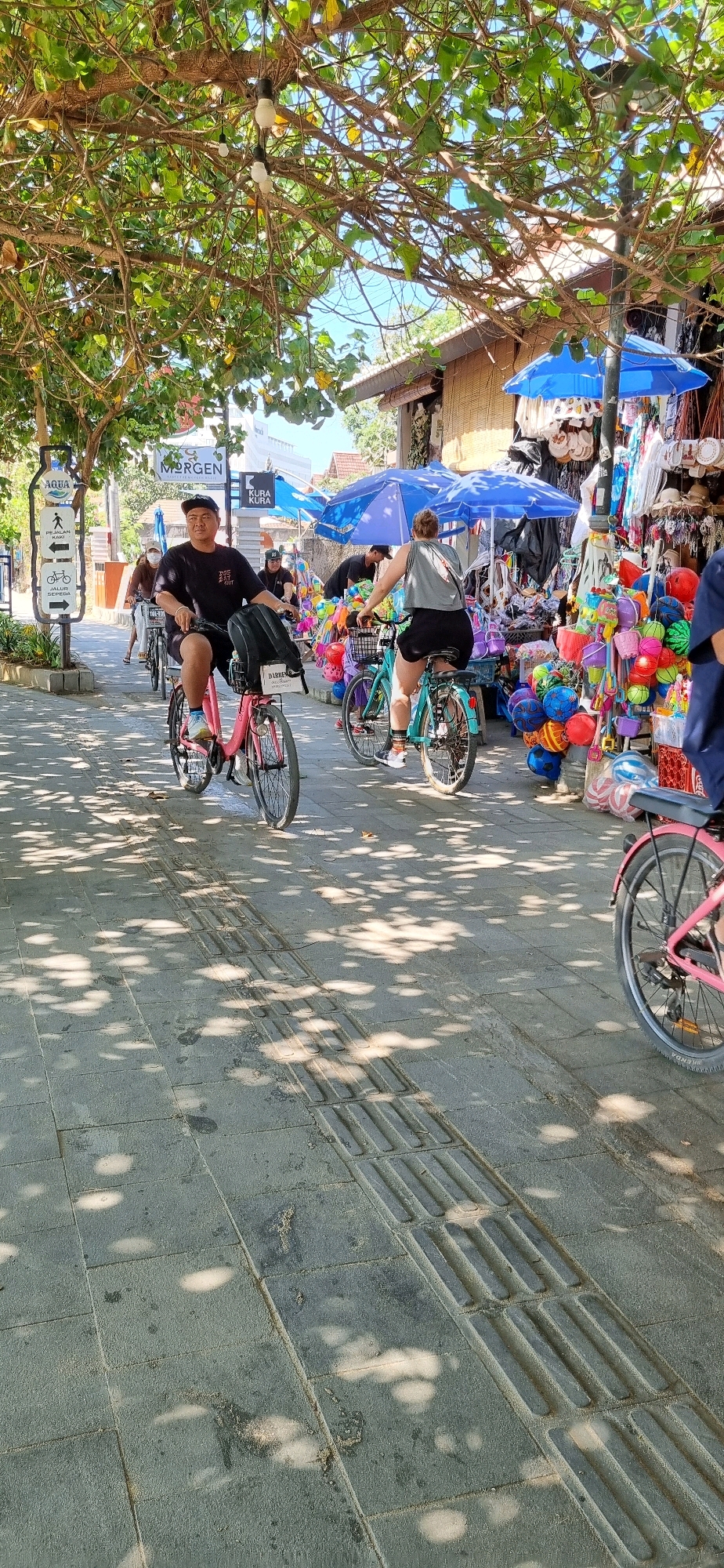 The image depicts a street scene in Denpasar, Indonesia, likely a tourist area given the presence of souvenir shops and people on bicycles. The foreground shows a textured gray paved sidewalk with tactile paving for the visually impaired. Sunlight filters through the canopy of a large tree, casting dappled shadows on the ground.
In the midground, several individuals are riding bicycles. A man in a black t-shirt and black shorts is on a pink bicycle, wearing a backpack. Another person, wearing a grey tank top and black shorts, is on a turquoise bicycle. Behind them, other cyclists are visible, as well as a vendor's stall overflowing with colorful goods, including numerous balls of various sizes and colors, hanging items, and blue umbrellas providing shade.
The background reveals buildings with woven-like facades, typical of Indonesian architecture, and more shops displaying merchandise. Signs such as "AQUA," "MORGEN," and "KURA KURA" are visible, suggesting businesses and potential tourist attractions. The overall atmosphere appears to be a sunny day in a lively urban environment.