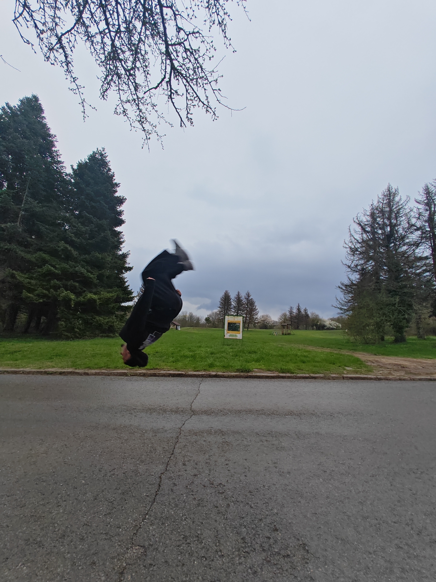 A person wearing dark clothing is captured mid-flip near a road in what appears to be a park or green space. The location is identifiable as Shumen, Bulgaria, due to the presence of a sign indicating a park area. The scene includes trees of different types, a grassy area, and a cloudy sky. The road in the foreground shows signs of wear with a visible crack.