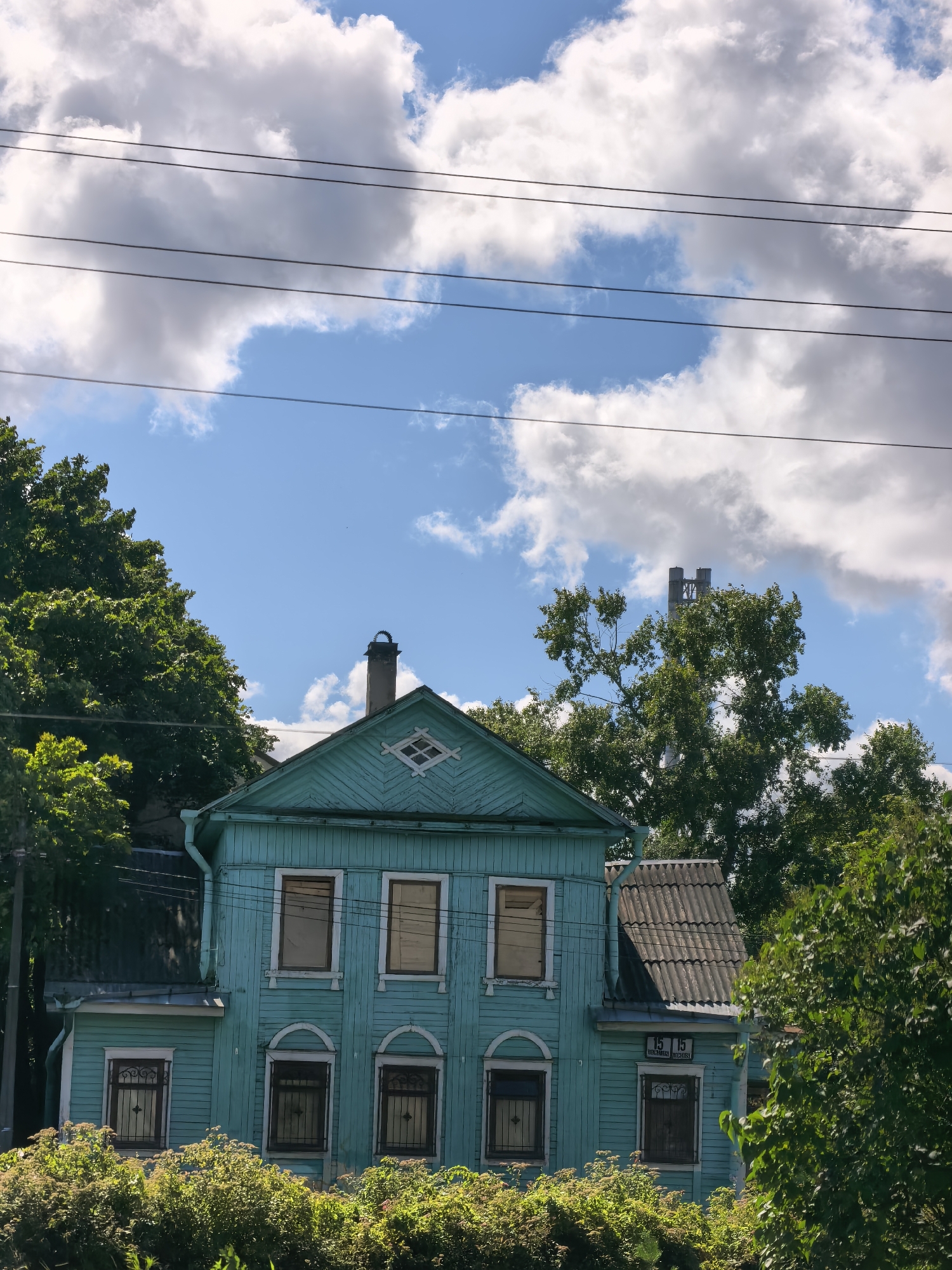 This is a medium shot, taken from a slightly low angle, of a turquoise, two-story wooden house with white trim in Sestroretsk, Russia. The house has a gable roof with gray shingles, and a chimney with a circular element on top. There are multiple windows on both floors, some with ornate black iron bars. The house is surrounded by lush green trees and foliage. In the background, there is a bright blue sky with scattered white clouds, and several power lines are visible stretching across the frame. In the distance, beyond the trees, two large industrial chimneys can be seen. The overall impression is of a slightly weathered but well-maintained residential building in a natural, somewhat overgrown setting, under a dynamic sky.