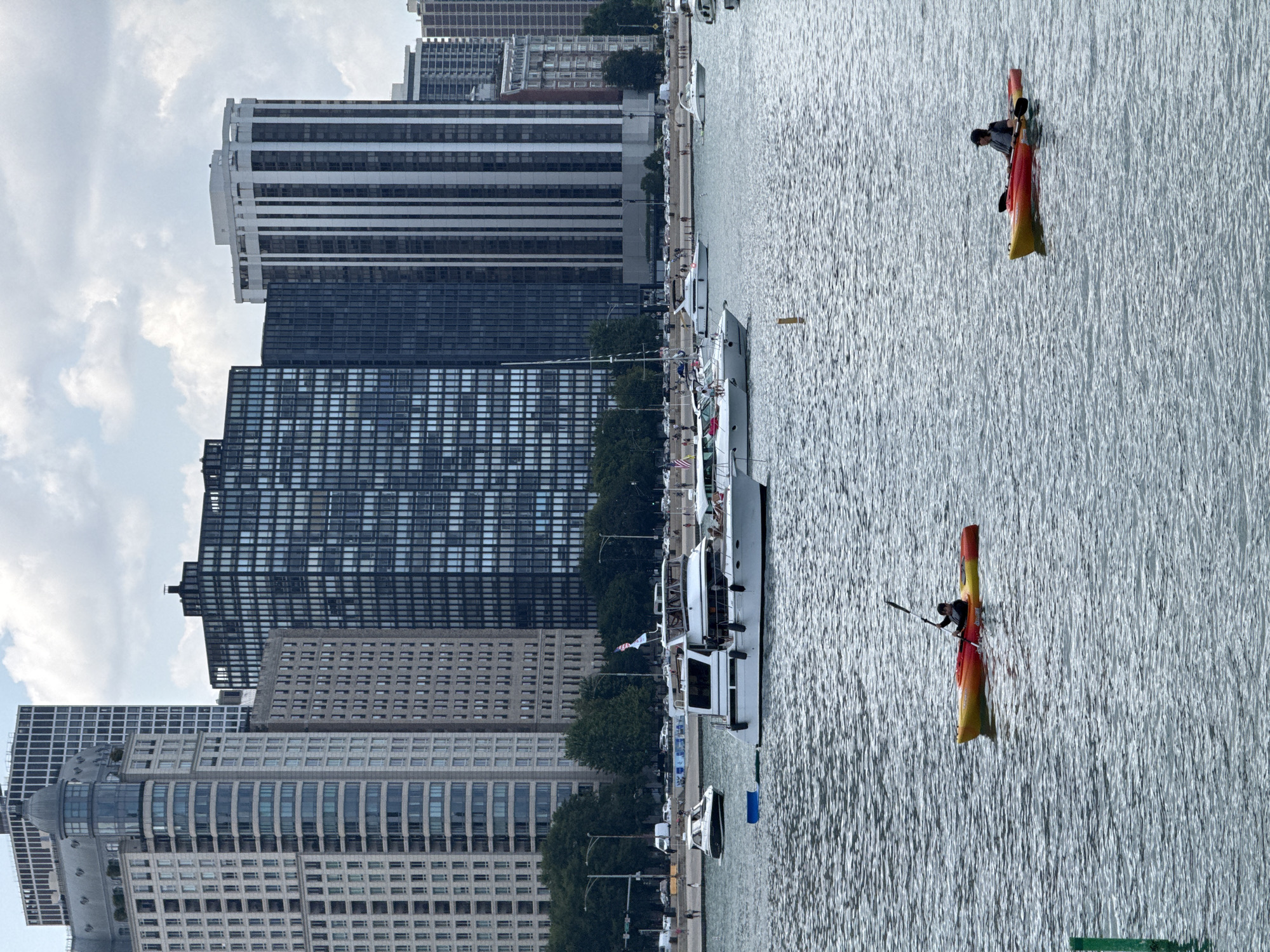 This image captures a view of Lake Michigan with the Chicago skyline in the background. The skyline features several modern high-rise buildings, including the distinctive rounded dome of a building on the left and a tall building with vertical white stripes on the right. In the foreground, the lake is dotted with various boats. A white yacht is prominently featured, with people visible on its deck. Several other smaller boats and a sailboat are also present. In the middle ground, two individuals are kayaking. One kayaker is closer to the viewer and is paddling a yellow and red kayak. Further to the right, another kayaker is paddling a similar colored kayak. The water is slightly rippled, reflecting the overcast sky. Trees and a shoreline with a road and people are visible behind the boats.