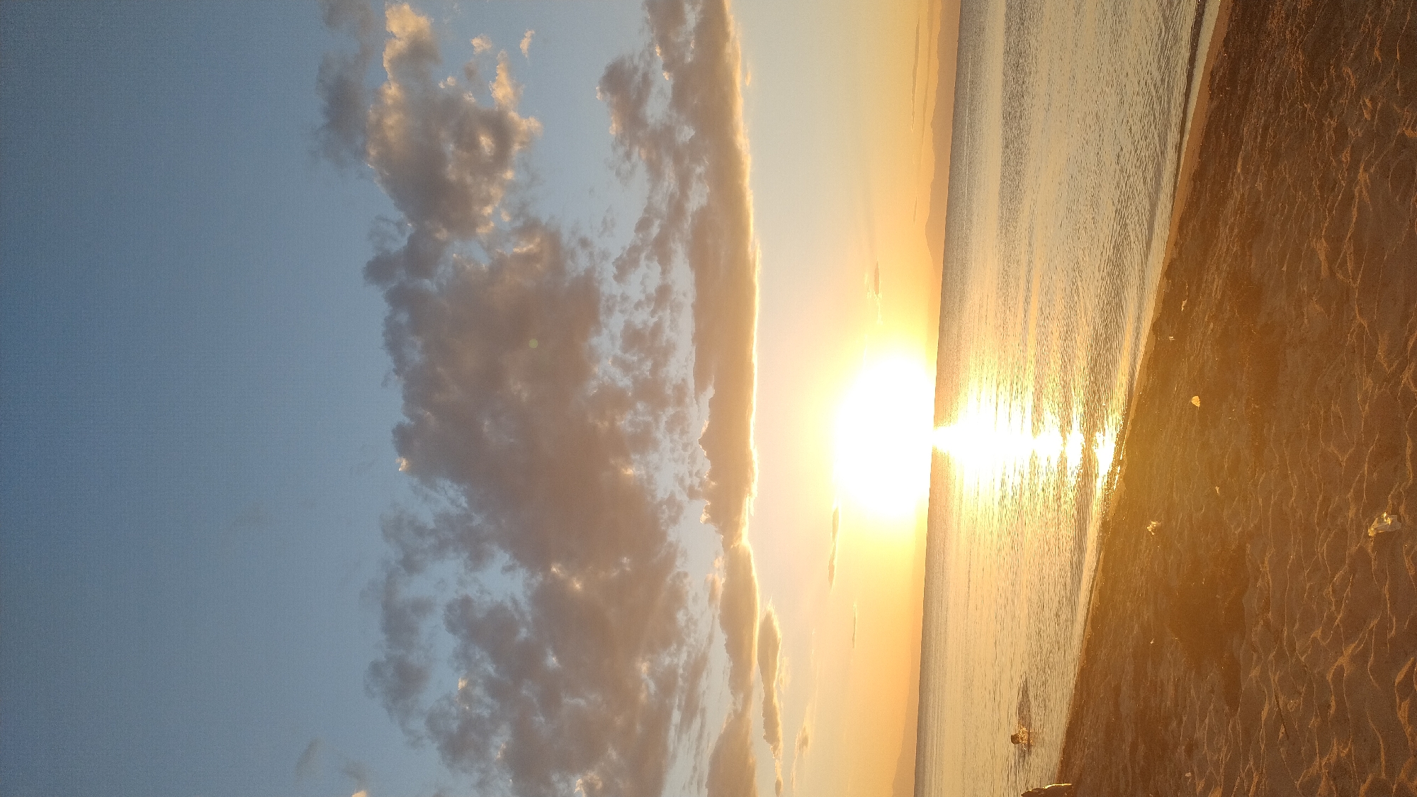 The image depicts a sunset over the sea at Perea, Greece. The sky is a gradient of blue and orange, with scattered clouds catching the sunlight. The sun itself is a bright, glowing orb on the horizon, casting a shimmering reflection on the calm water. A sandy beach occupies the foreground, with some plastic debris visible on the sand. In the distance, a faint outline of mountains can be seen on the horizon. Two individuals are present in the water, one of whom appears to be swimming. The overall scene conveys a tranquil and serene atmosphere.