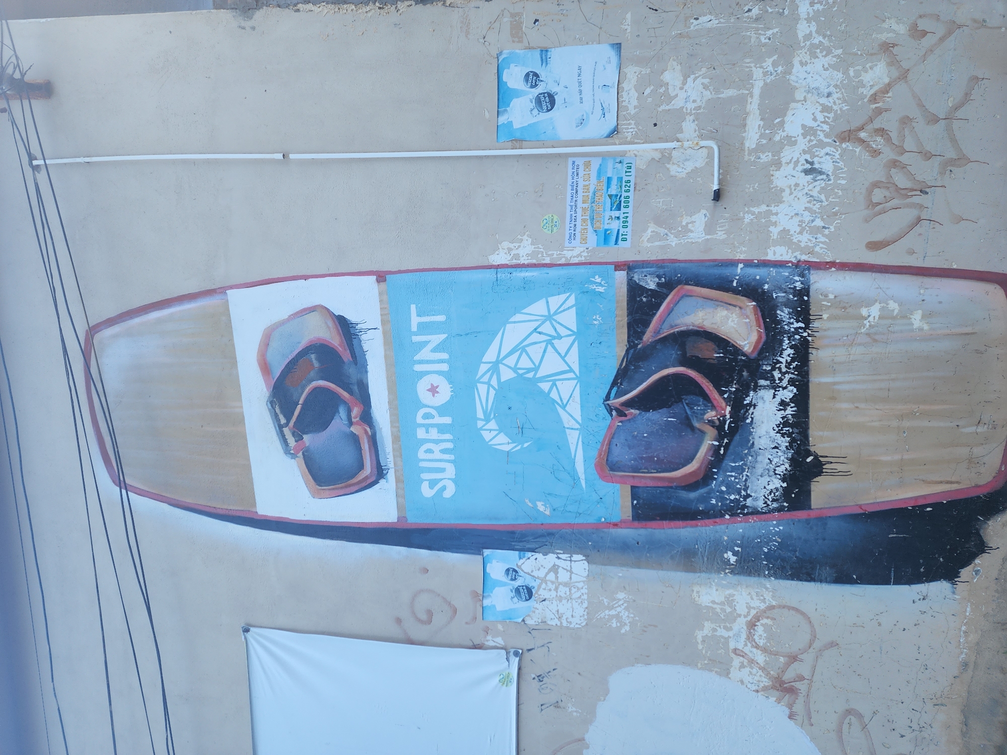 The image depicts a wall with graffiti art in Phu Thuy Ward, Vietnam. The central element is a large mural of a surfboard, rendered in a stylized manner with a prominent wave design and what appears to be a pair of sunglasses. The surfboard art is vibrant, with sections of blue, white, and brown. Below the surfboard mural, there are various posters and stickers affixed to the wall, some of which are in Vietnamese. One poster advertises a sports company with contact information. There is also a white banner hanging on the left side of the wall. The wall itself shows signs of wear and tear, with peeling paint and some additional graffiti. The environment appears to be an outdoor urban setting, with overhead wires visible, suggesting it is part of a building's exterior. There are no people or animals present in the image.