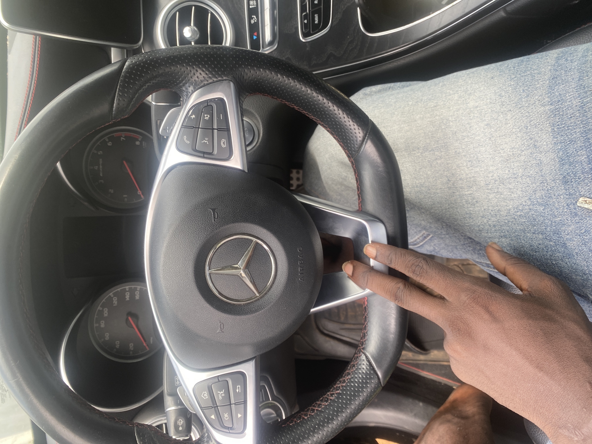 The image shows the interior of a Mercedes-Benz car. The steering wheel is visible, with the Mercedes-Benz logo at its center. The dashboard has a speedometer that reads approximately 100 mph. A person's hands are visible, one resting on the steering wheel and the other extended towards the camera. The person is wearing light blue jeans. The car's interior appears to be in good condition. The location is identified as Oniyangi, Nigeria, though there is no visual evidence in the image to confirm this specific location.