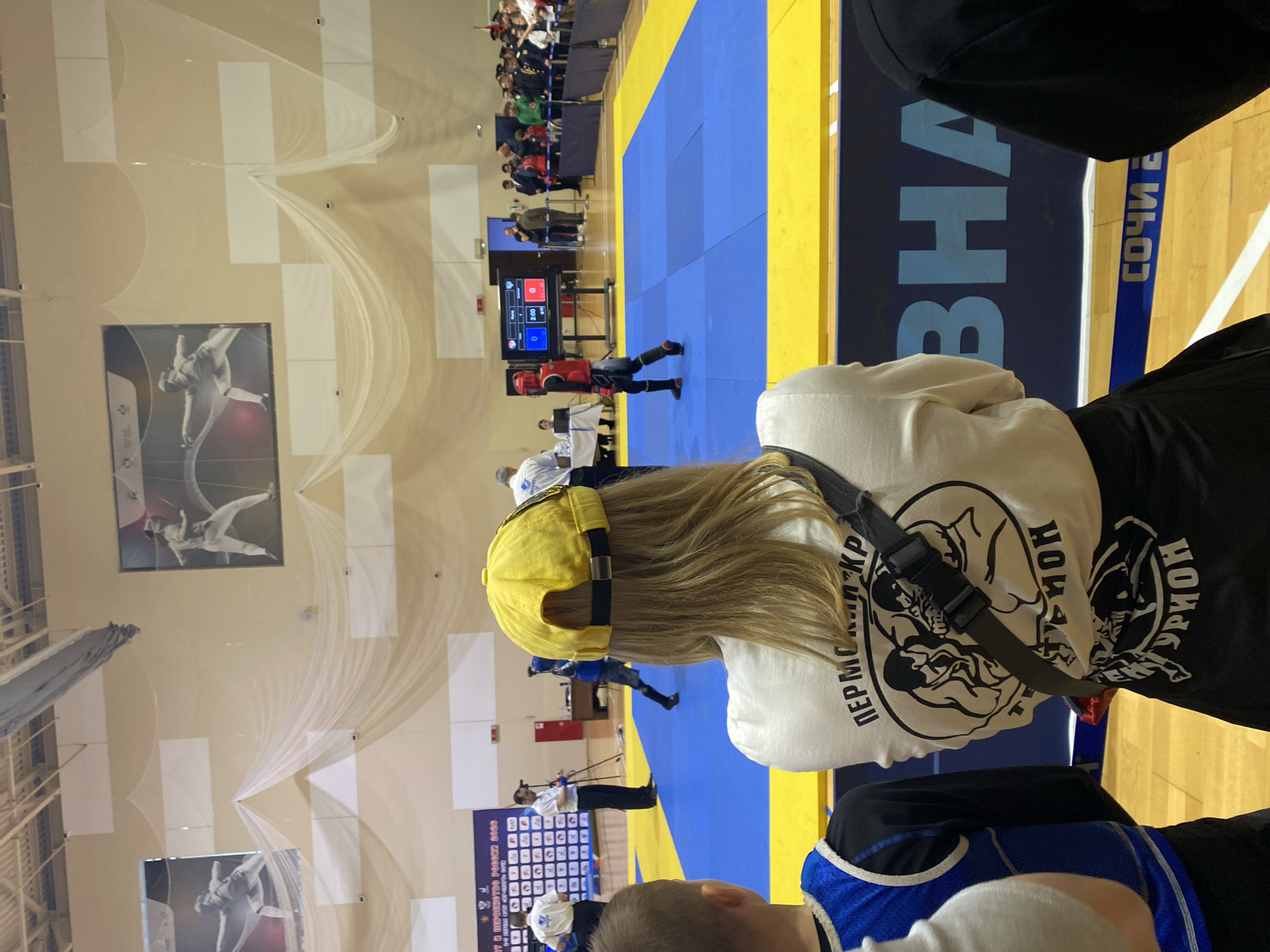 A spectator views a combat sports event in Sochi, Russia, as evidenced by a sign in the background. A blue and yellow mat is visible where the matches are being held. There are other spectators present in the arena. Two rectangular framed art pieces are displayed on the wall behind the mat. A woman in the foreground wears a yellow baseball cap and a white shirt. A black strap extends across her body. The image is taken from a close perspective, focusing on the woman.