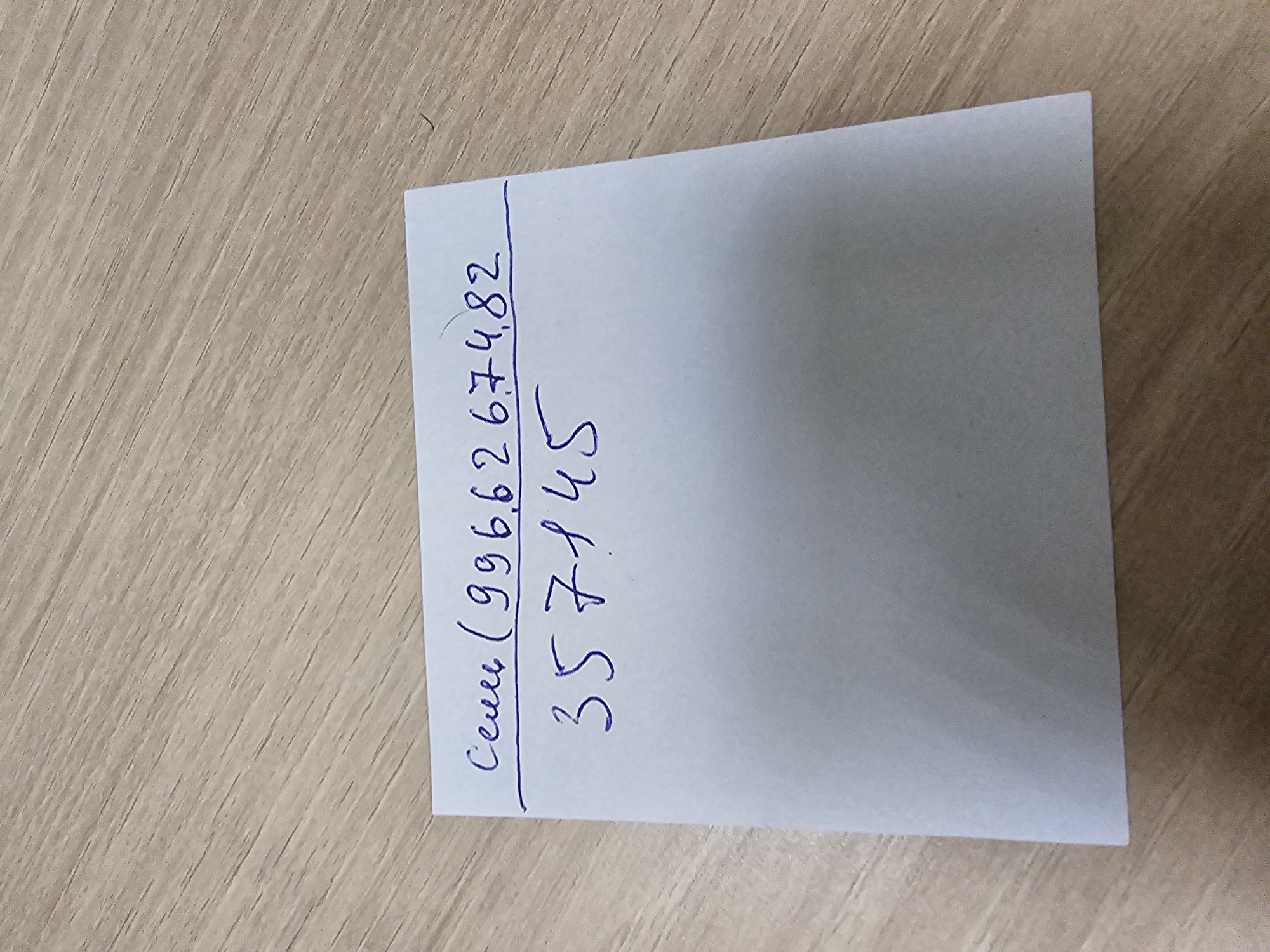 A piece of white paper with handwriting in blue ink lies on a light-colored wooden surface. The handwriting on the paper consists of two lines. The top line reads "сени (996,6267,4,82", and the bottom line reads "357145". The paper is positioned at an angle. The wooden surface has a prominent grain pattern. There are no people visible in the image. The scene appears to be indoors.