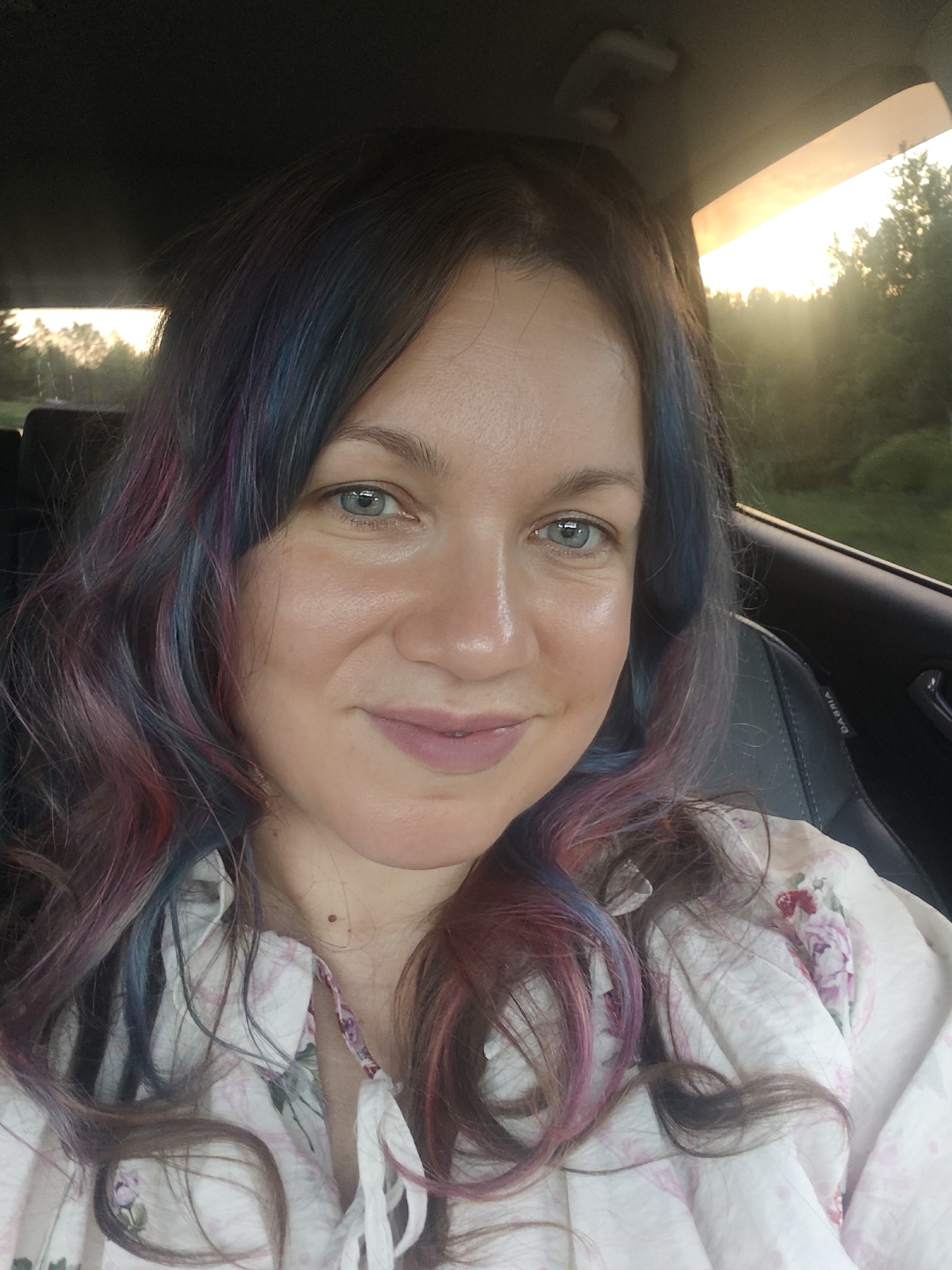 A selfie of a woman with fair skin, blue eyes, and multicolored hair (dark brown with streaks of blue, red, and pink). She is wearing a light-colored floral print blouse. The background appears to be the interior of a car, with a glimpse of a wooded area and a sunset through the window. It is not possible to confirm if it is in the city of Ильинское, Russia.