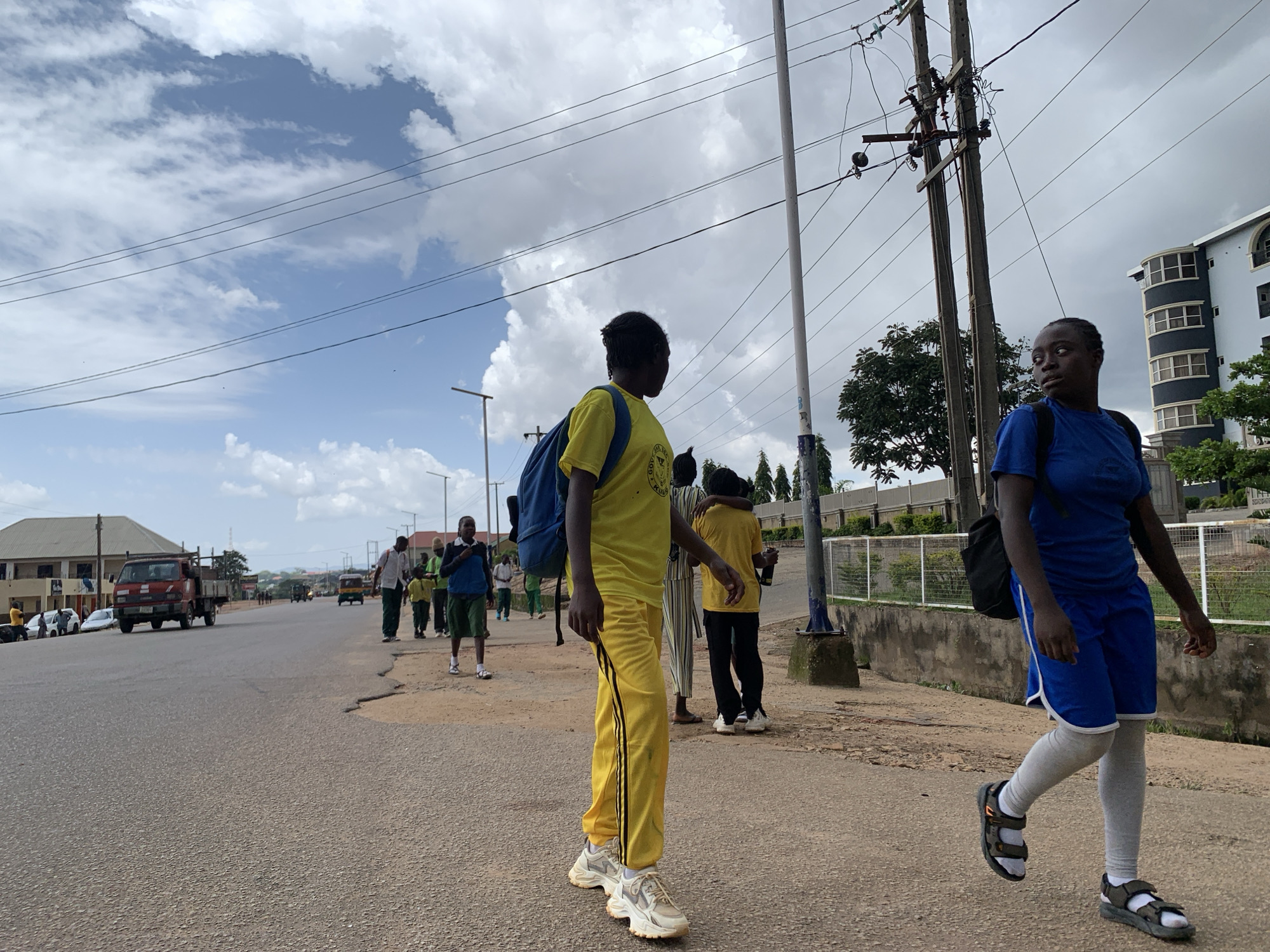 The image depicts a street scene in Jos, Nigeria, under a cloudy sky. The foreground features two young people walking on a paved road. The person on the left, facing away from the camera, wears a bright yellow t-shirt and matching yellow pants with black stripes, carrying a blue backpack. The person on the right, also facing away from the camera, is dressed in a blue t-shirt, blue shorts with white trim, and grey leg warmers, wearing sandals.
In the background, several other individuals, likely students, are walking along the side of the road, some carrying backpacks. A red truck is visible on the road, as are other vehicles and buildings, including a modern-looking, multi-story building with rounded balconies. Power lines and utility poles are prominent features of the urban landscape. The weather appears overcast, suggesting it might be daytime, but the exact time is not discernible. The overall atmosphere suggests a typical day with people commuting or moving about their daily activities in an urban environment. There is no visible text in the image.