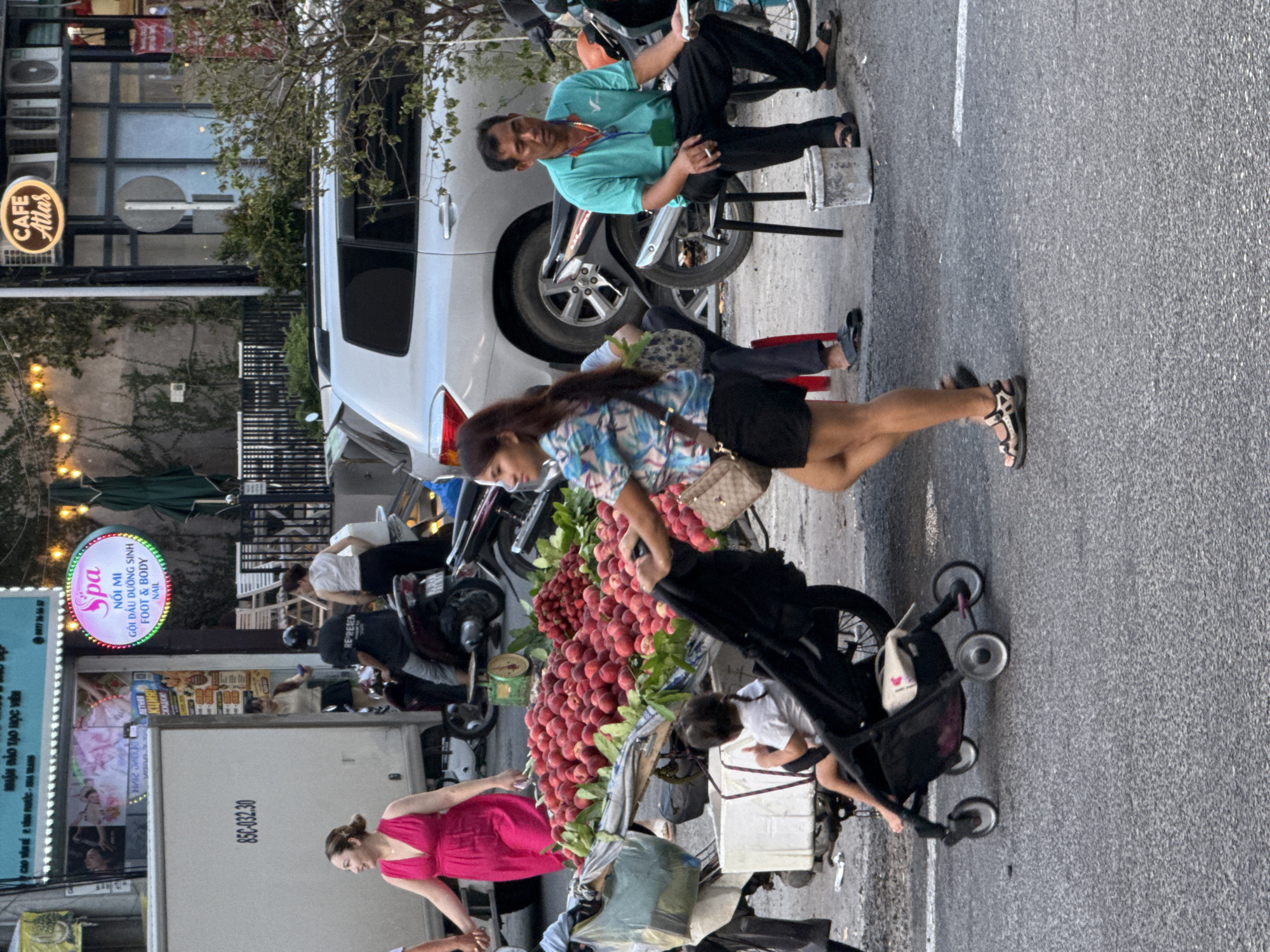 The image depicts a street scene in Bac Nha Trang Ward, Vietnam, at what appears to be dusk. In the foreground, a woman in a patterned shirt and black shorts pushes a stroller containing a young child. To her right, a man in a teal shirt and black pants sits on a stool, holding a cigarette and a small item, possibly cash. Behind the woman and stroller, a cart laden with a large pile of red fruits, possibly peaches or nectarines, is visible. A white truck with the license plate "85C-032.30" is parked to the left. Further back, several motorcycles are parked, and a silver SUV is visible. A building with signage that includes "CAFE Atlas" and a spa advertisement with the text "SPA NÔI MÌ FOOT & BODY NAIL" is present in the background, illuminated by string lights. The ground is paved asphalt, and the overall setting suggests a bustling market or roadside vendor area.