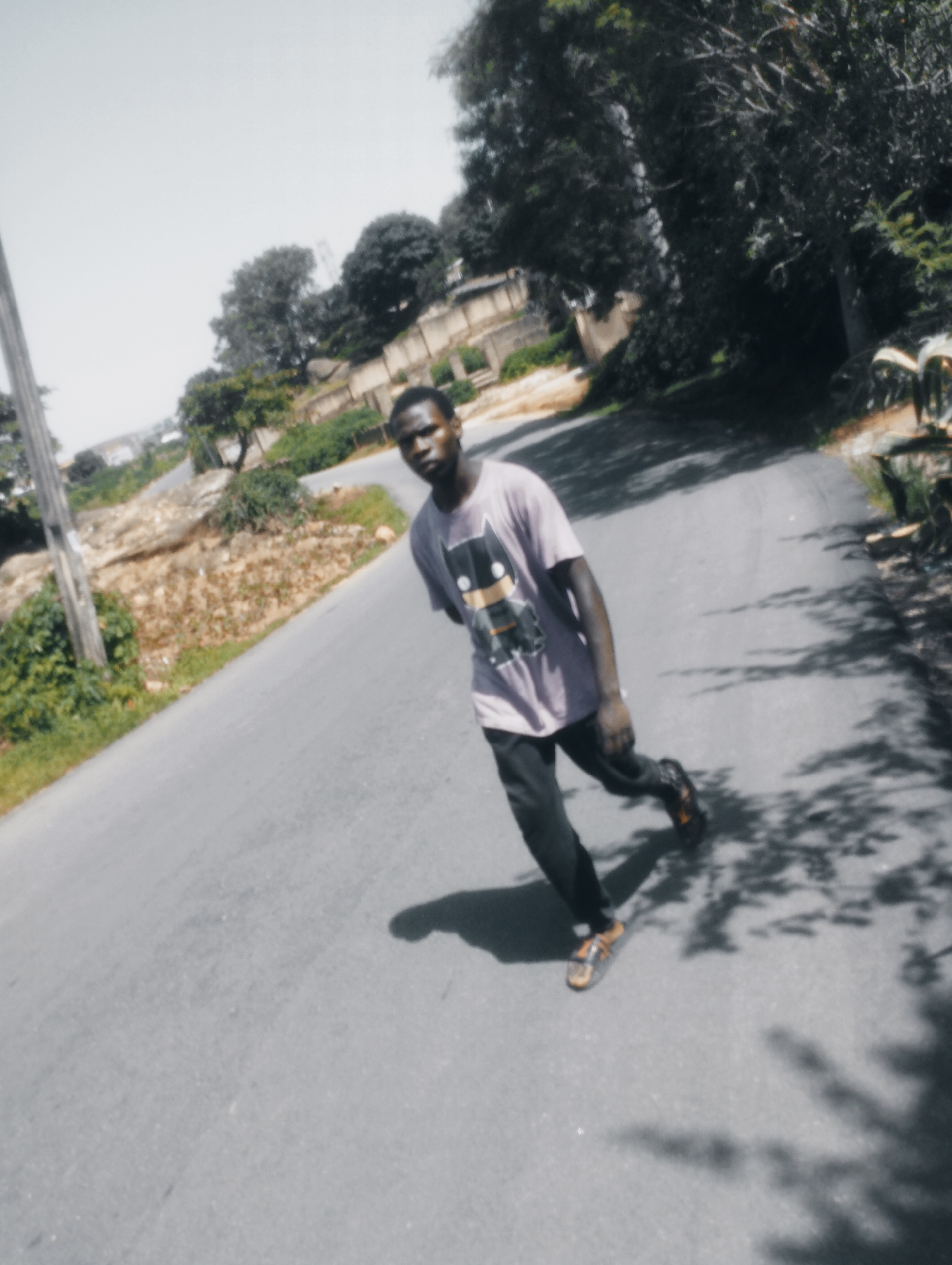 A young man with dark skin and short hair is captured in a dynamic pose on an asphalt road. He is wearing a light purple t-shirt with a graphic design of what appears to be the character Batman. His t-shirt is paired with dark pants and brown sandals. The man is facing slightly away from the camera and appears to be mid-stride, leaning into a turn. The road is an empty, paved surface with a faint yellow line marking its edge on the left. To the left of the road, there is a steep, rocky incline covered in sparse vegetation and a weathered grey pole. Beyond this incline, there are buildings and trees. To the right of the road, dense foliage and trees cast dappled shadows on the asphalt. The sky is bright and overcast. This scene is consistent with a location in Jos, Nigeria, characterized by its hilly terrain and mix of urban development and natural vegetation.