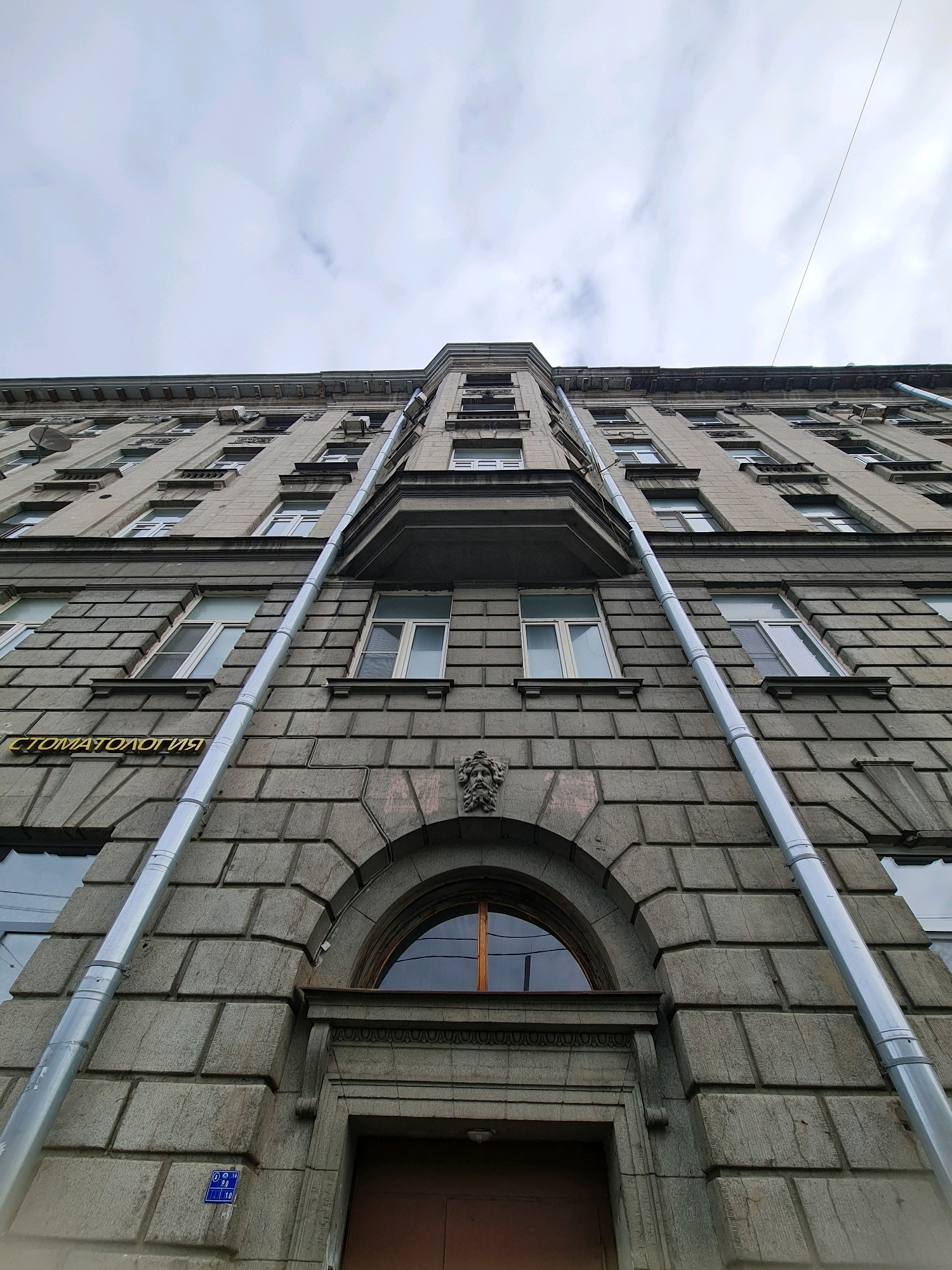 This is a low-angle, wide-shot photograph of the facade of an old building, likely in Saint Petersburg, Russia, based on the Cyrillic text on the building. The building is made of stone blocks and has numerous windows, some with white frames. Two large, silver downspouts run vertically along the facade, meeting at the bottom of the frame near an arched entryway. The sky above is overcast with clouds. A sign with Cyrillic text, reading "СТОМАТОЛОГИЯ," which translates to "DENTISTRY," is visible on the left side of the building. A small blue sign with white numbers is also present on the lower left.
