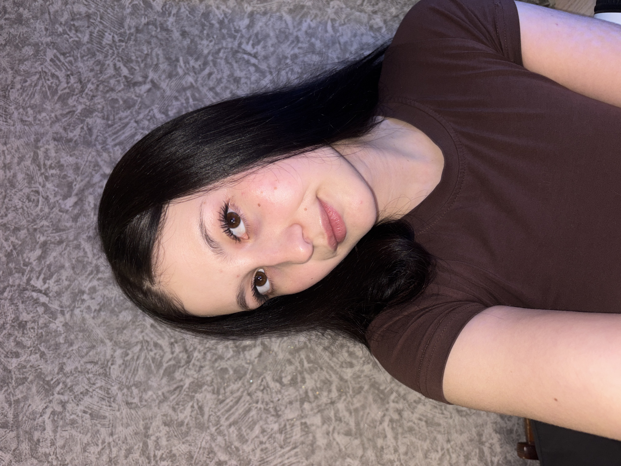 A woman with dark hair is lying on a gray textured surface. She is wearing a brown t-shirt. Her face is angled towards the camera. The location is indiscernible from the information in the image.