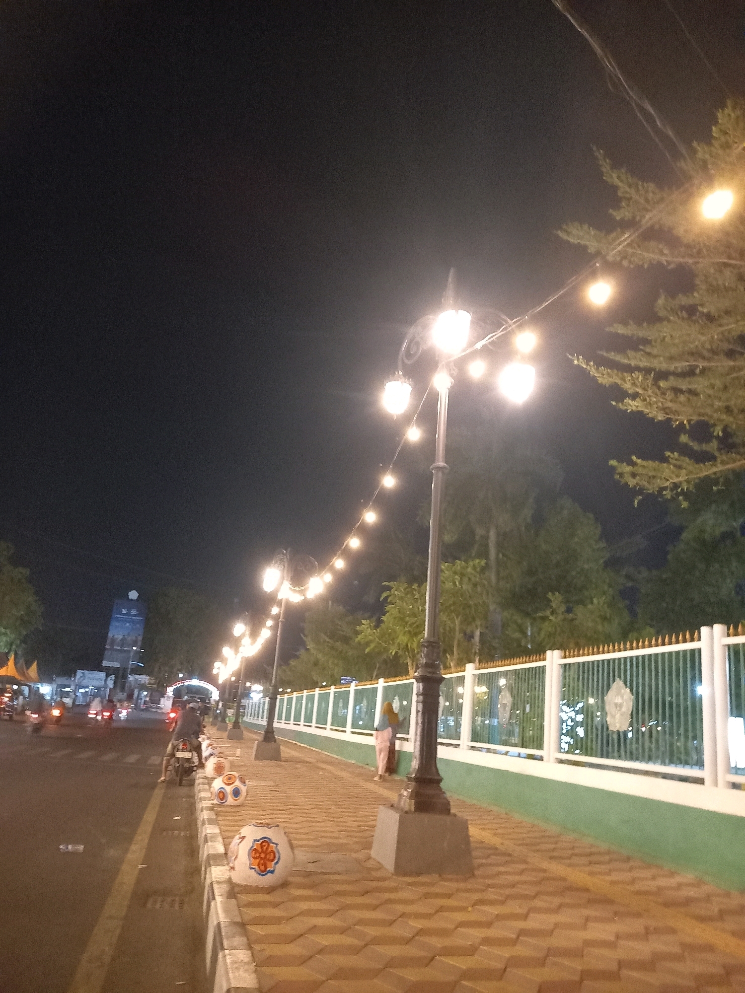 The image depicts a street scene at night in Pamekasan, Indonesia. A row of ornamental streetlights with warm-toned bulbs illuminates a sidewalk paved with brown interlocking bricks. The sidewalk is bordered by a low green wall topped with a white fence. Decorative spherical planters, some painted with orange and blue patterns, are placed along the sidewalk. On the street, several motorcycles are visible, with one rider heading away from the camera. The street itself is dark, with yellow lane markings. In the background, trees and buildings are dimly lit, and a tall, dark structure with a blue accent is visible in the distance. The overall atmosphere is that of a well-lit urban area at night.