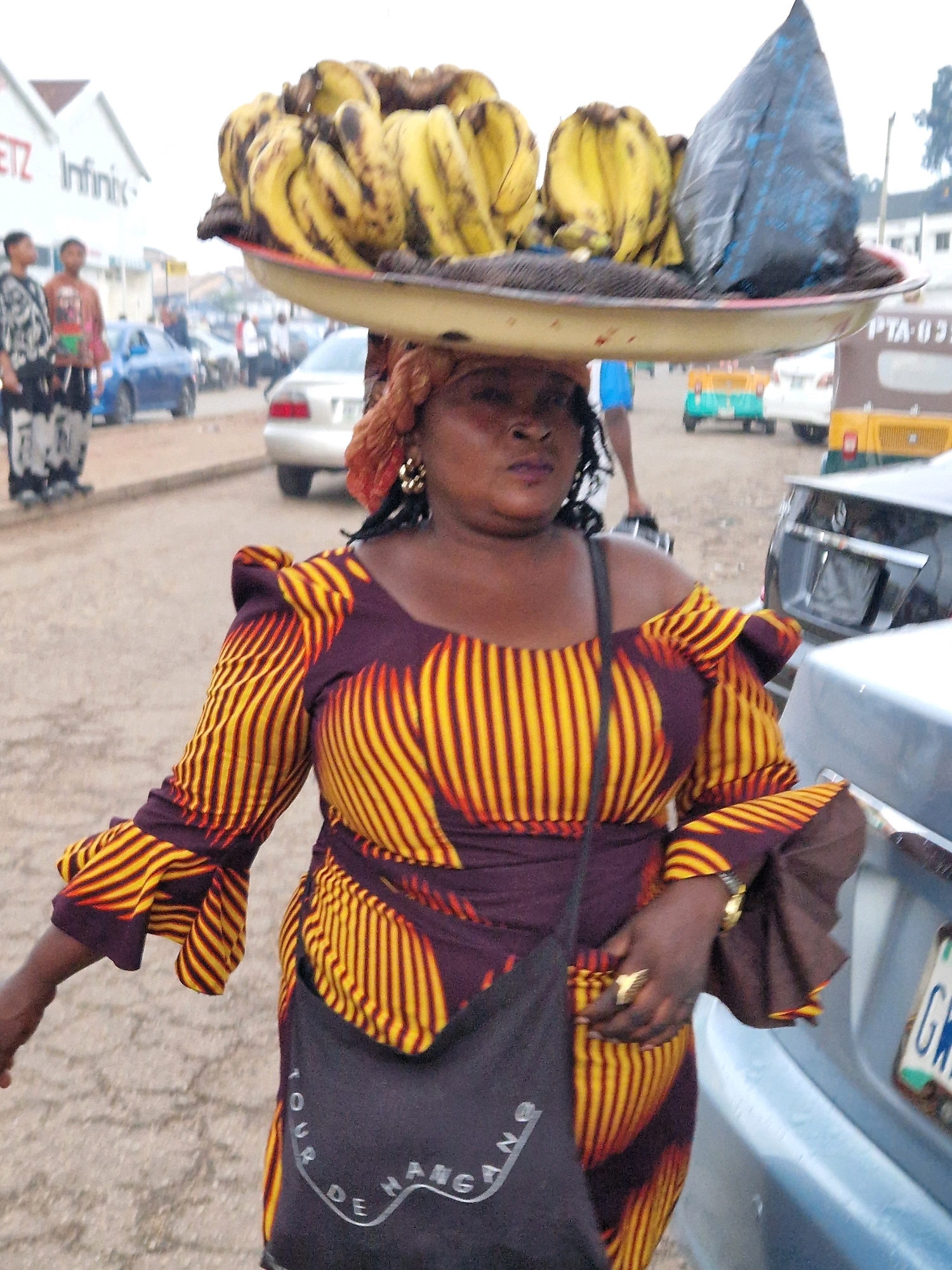 A dark-skinned woman, wearing a colorful patterned dress and gold earrings, carries a large tray of bananas and a black plastic bag balanced on her head. She walks down a paved street in Jos, Nigeria. A dark-colored car is parked next to her, and a license plate with the letters "GW" is visible. In the background, other cars and buildings can be seen. The woman has dark braided hair and is wearing a ring on her right hand. A black bag hangs from her shoulder.