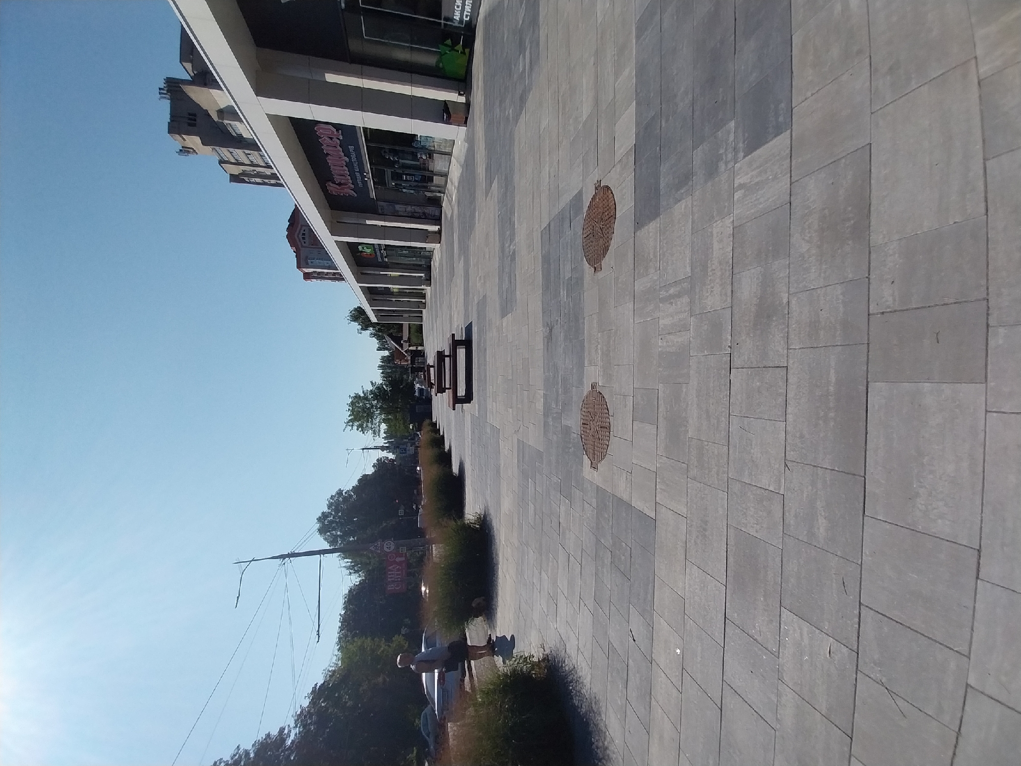 The image shows a modern pedestrian zone in the city of Dnipro, Ukraine, likely in front of a commercial building. The ground is paved with large, gray rectangular tiles laid in a pattern. Several wooden benches are arranged on the pavement, and there are rows of ornamental grasses planted in the foreground.
In the background, a long, white-colored building with a modern facade dominates the right side of the image. The building appears to be a commercial establishment with several storefronts, one of which has a sign with Cyrillic script that reads "Kupaper" and "ГУРТОВЫЙ КАНЦТОВАРІВ" (Wholesale Stationery). Above the building, a tall, multi-story residential building with visible architectural details can be seen against a clear blue sky.
On the left side of the image, a person wearing a light-colored t-shirt and dark shorts stands near a cluster of bushes, facing away from the camera. Cars are visible on a street to the left, partially obscured by trees and bushes. A street light and power lines are also present in the background. Two circular manhole covers are visible on the pavement. The overall impression is of a well-maintained urban public space on a sunny day.