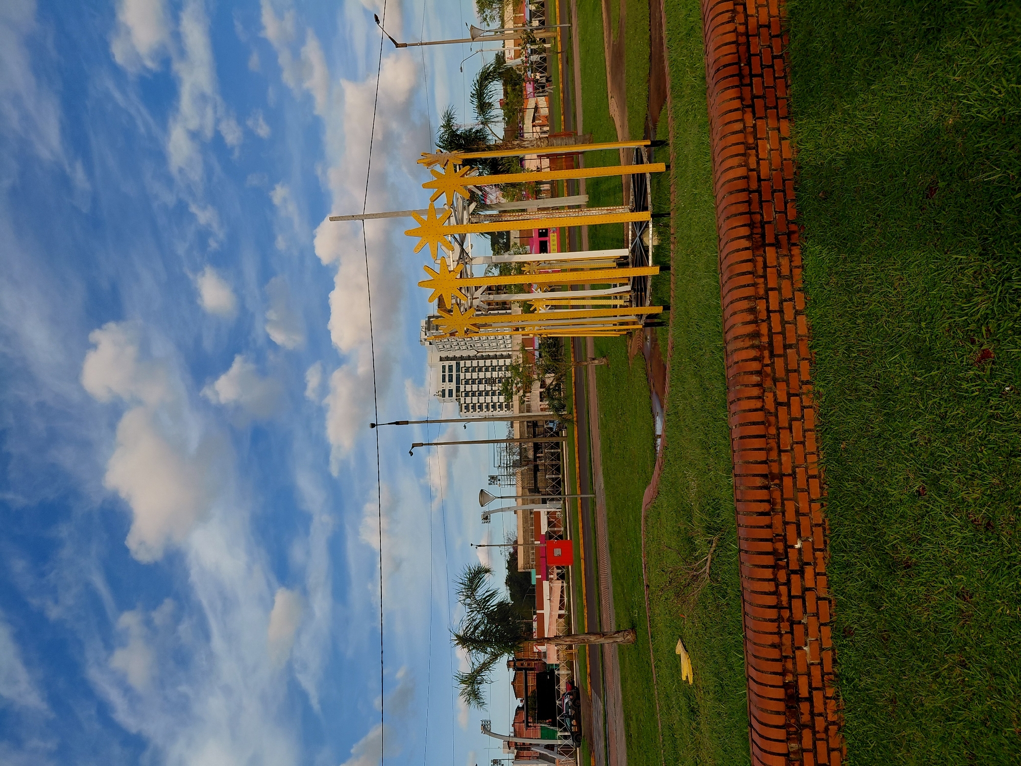 The image depicts an outdoor scene in Encarnacion, Paraguay, likely in a park or public space. In the foreground, a low, curved brick wall separates a patch of grass from a wider expanse of green lawn. A yellow, star-shaped structure, possibly part of a playground or decorative element, stands on the lawn behind the brick wall. The middle ground features a paved road with a traffic light and a few palm trees. Beyond the road, buildings of varying heights are visible, including a tall, modern, multi-story building and other structures that appear to be commercial or recreational. In the background, a bright blue sky with scattered white clouds completes the scene. The overall impression is one of a well-maintained urban area on a clear day. No people are visible in the image.